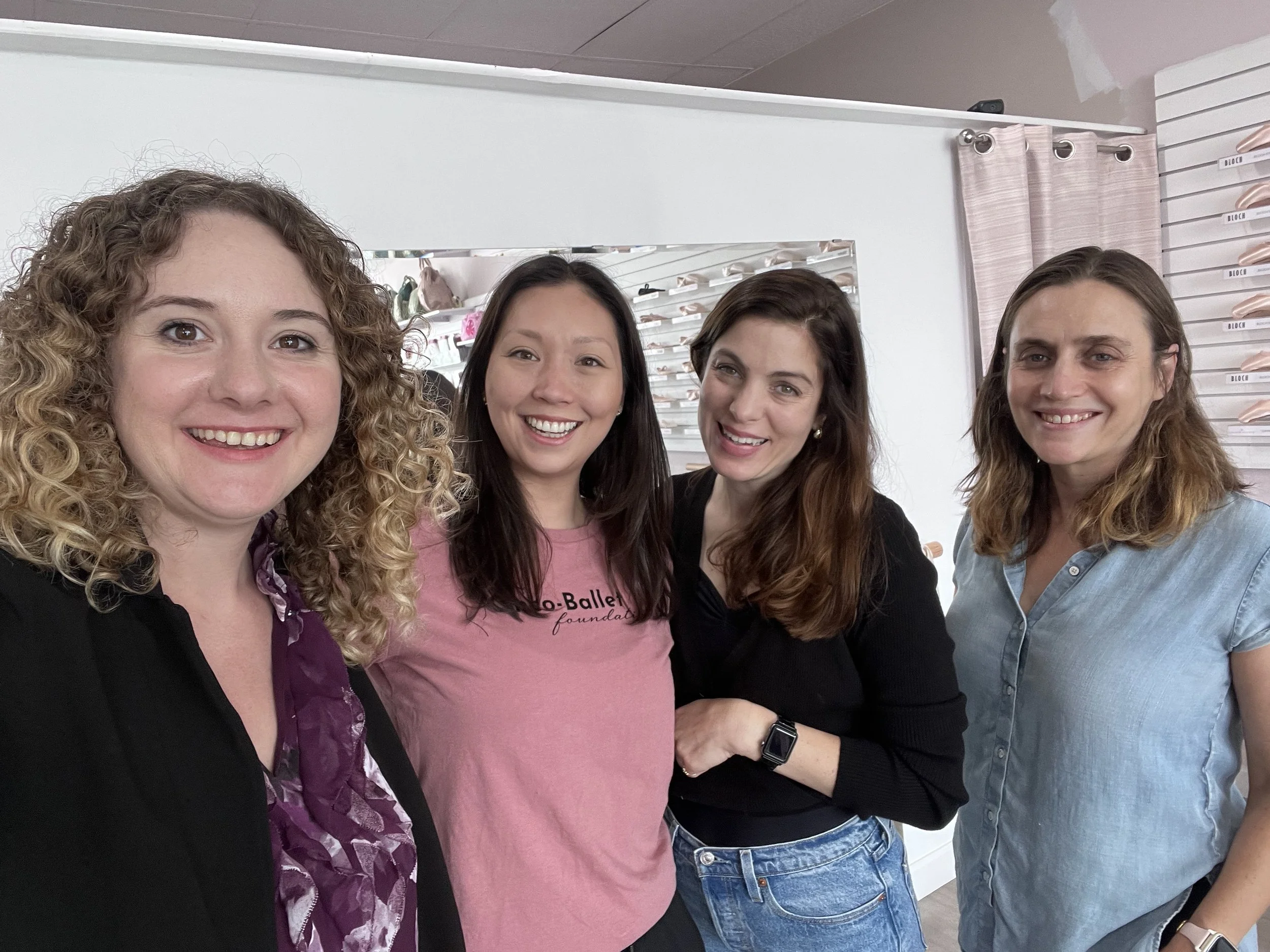 Four women smiling for a group photo in an indoor setting, with shoes displayed on wall shelves behind them.
