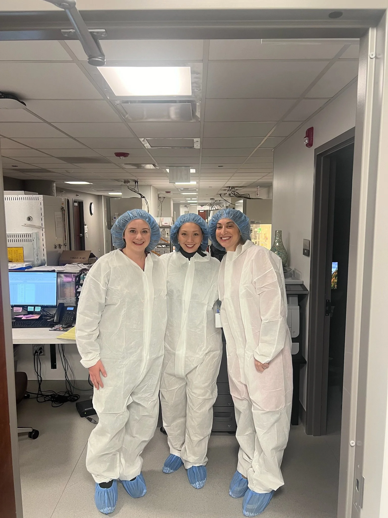 Three women in protective medical gowns, hair caps, shoe covers, and gloves standing in a hospital or laboratory corridor.