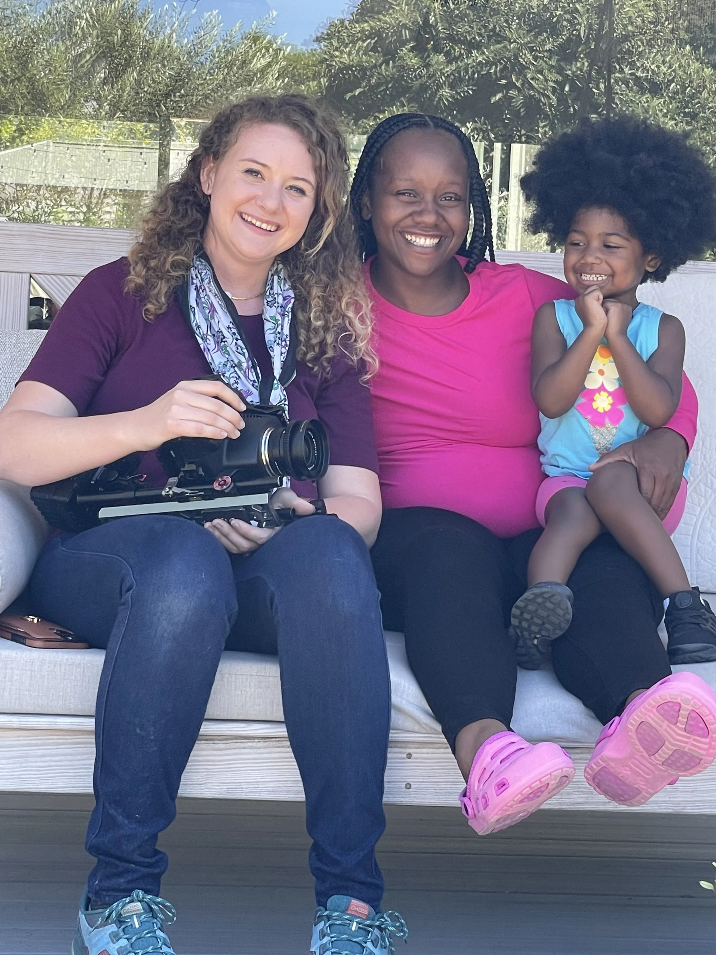 Three women sitting on a white outdoor couch, smiling and posing for a photo. One woman is holding a camera. Two women have dark curly hair, one with long braids, and the third woman has light brown, curly hair. They are dressed casually, with the wo