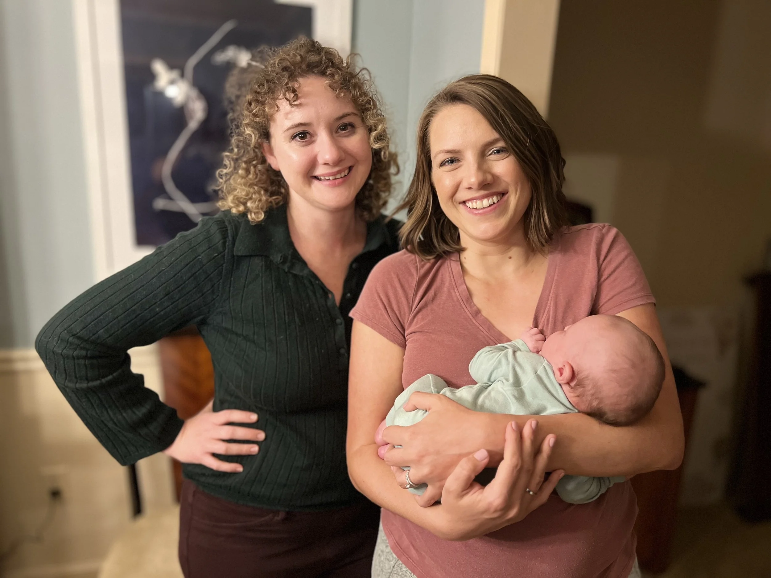Two smiling women, one with curly hair and the other with straight hair, and a baby wrapped in a blanket.