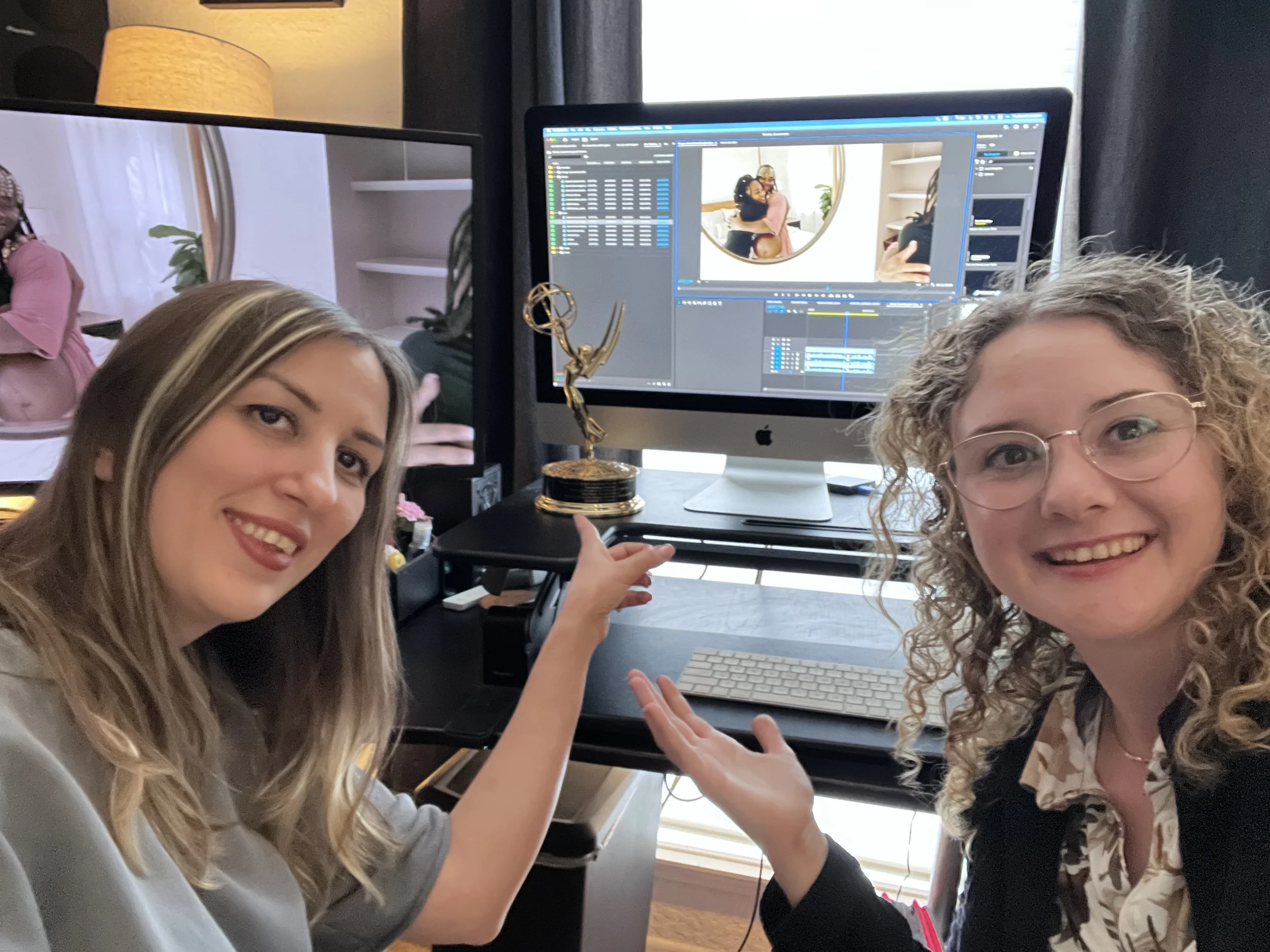 Two women are smiling and posing in front of computer monitors showing photo editing software. One woman has long blond hair, and the other has curly blond hair and glasses. They are inside a room with a window and some shelves behind them.