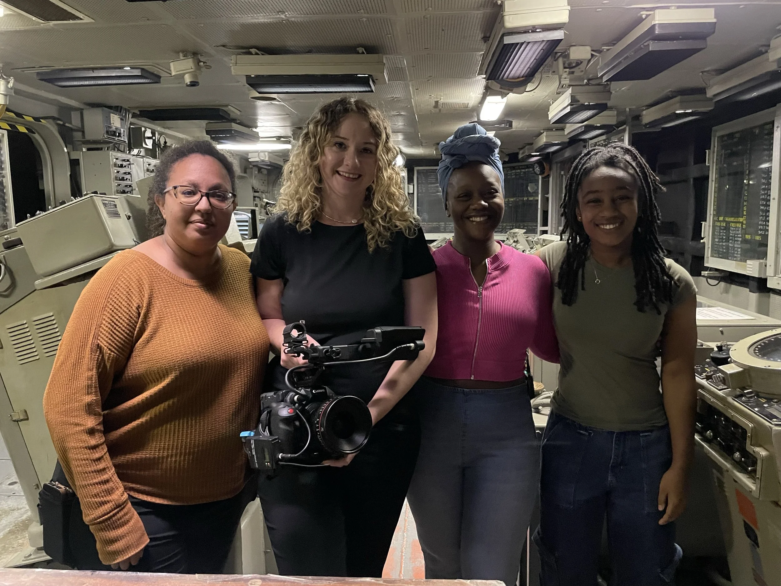 Four women standing together in a scientific or technical environment with machinery and equipment behind them.
