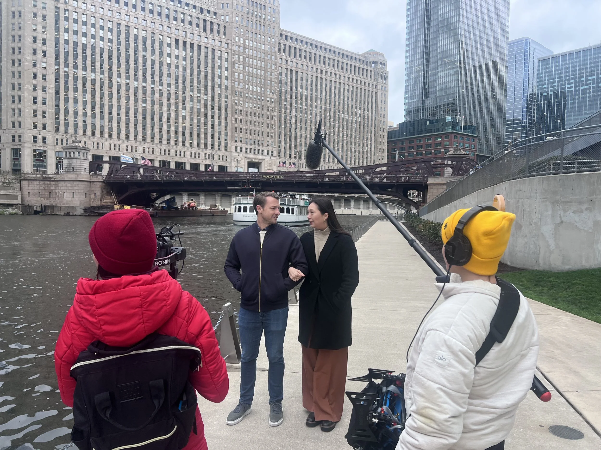 Two people being interviewed outdoors by a camera crew, with tall city buildings and a river in the background.