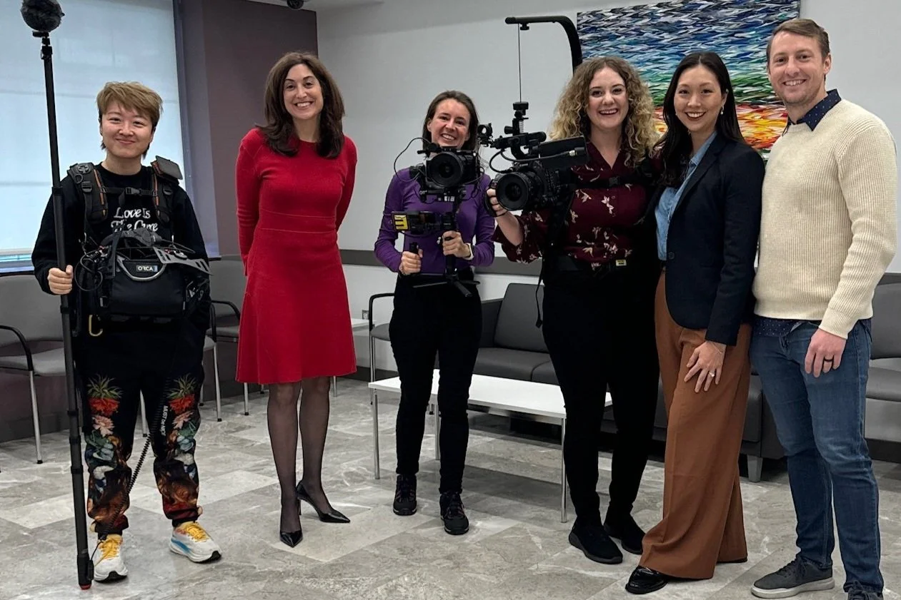 Six people standing together in an indoor setting, two holding cameras with stabilization equipment, and one with crutches. They are smiling and dressed in casual and business attire.