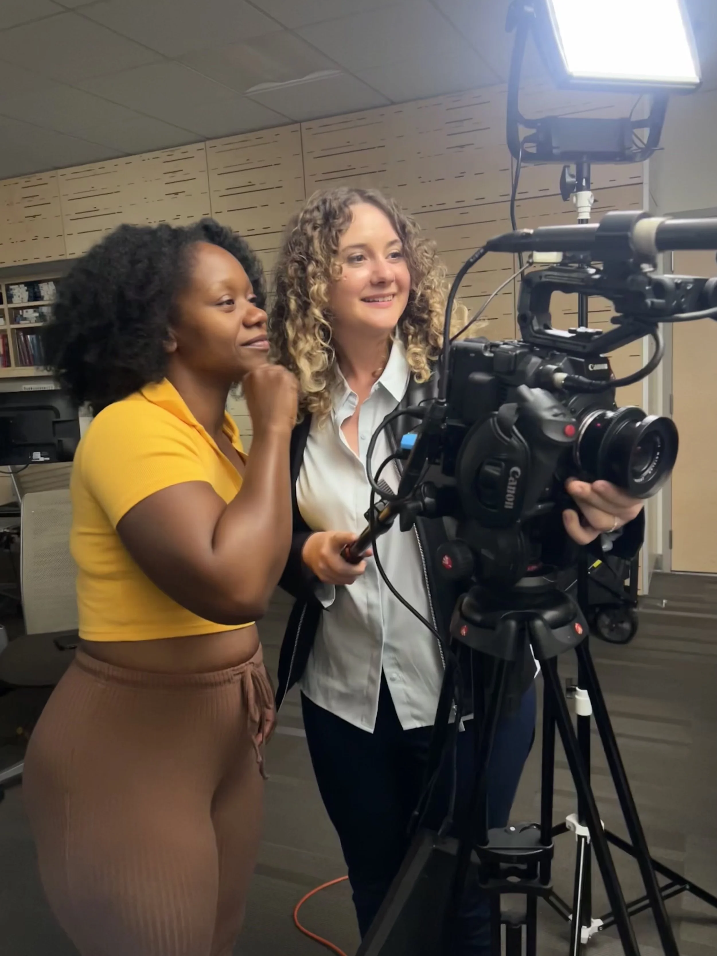 Two women operating a professional video camera in an indoor setting, with one woman in a yellow shirt and the other in a white shirt with curly hair, both focused on filming or reviewing footage.