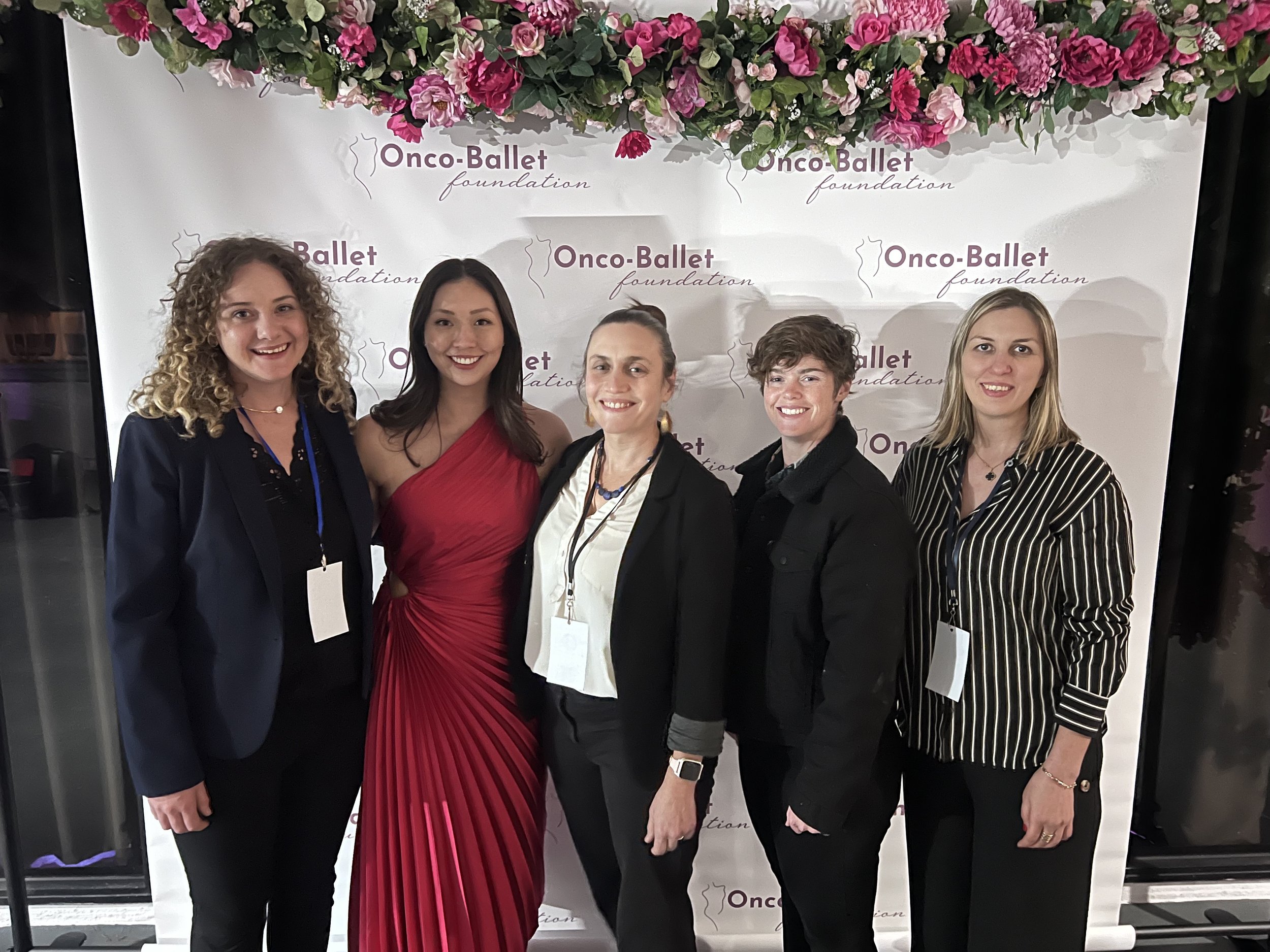 Five women standing in front of a backdrop with pink flowers and 'Onco-Ballet foundation' written on it. They are dressed in formal attire and smiling at the camera.