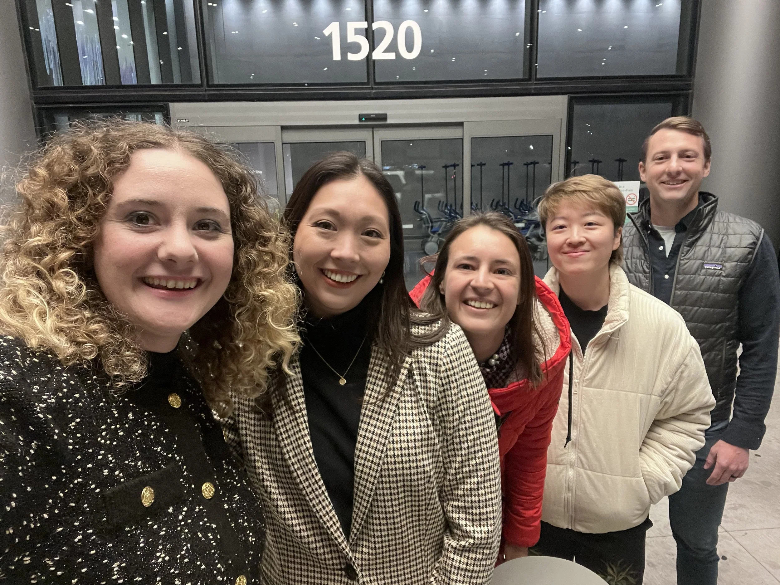 Group of six people smiling, standing in front of a glass window with the number 1520 above it, inside an airport terminal.