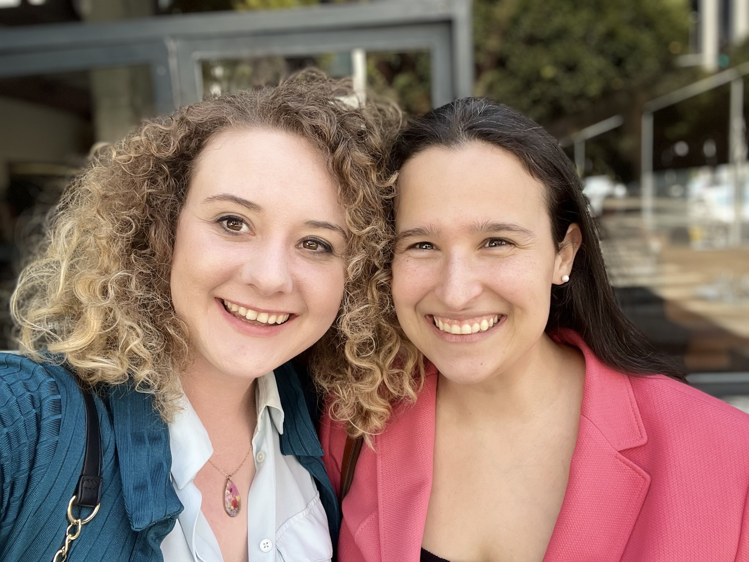 Close-up of two women smiling, with one having curly blonde hair and the other with straight dark hair, outdoors with trees and a metal structure in the background.