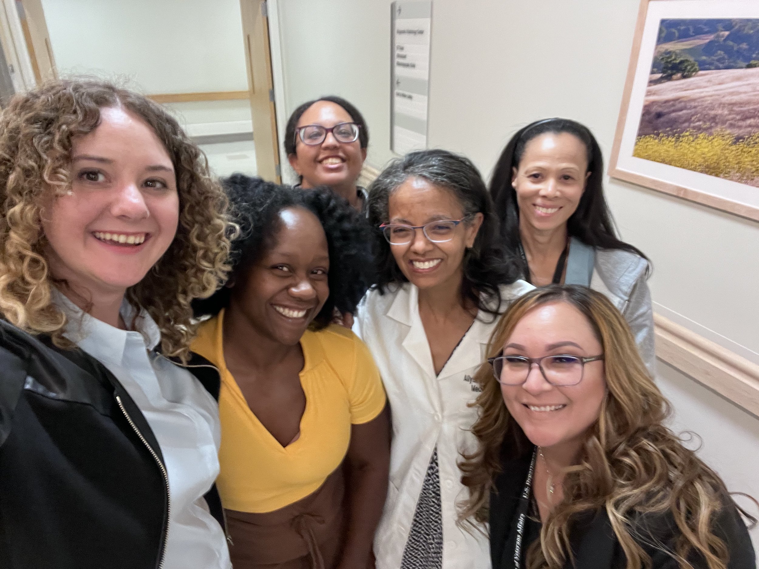 A group of women smiling taking a selfie in an indoor setting with a framed landscape photograph on the wall behind them.