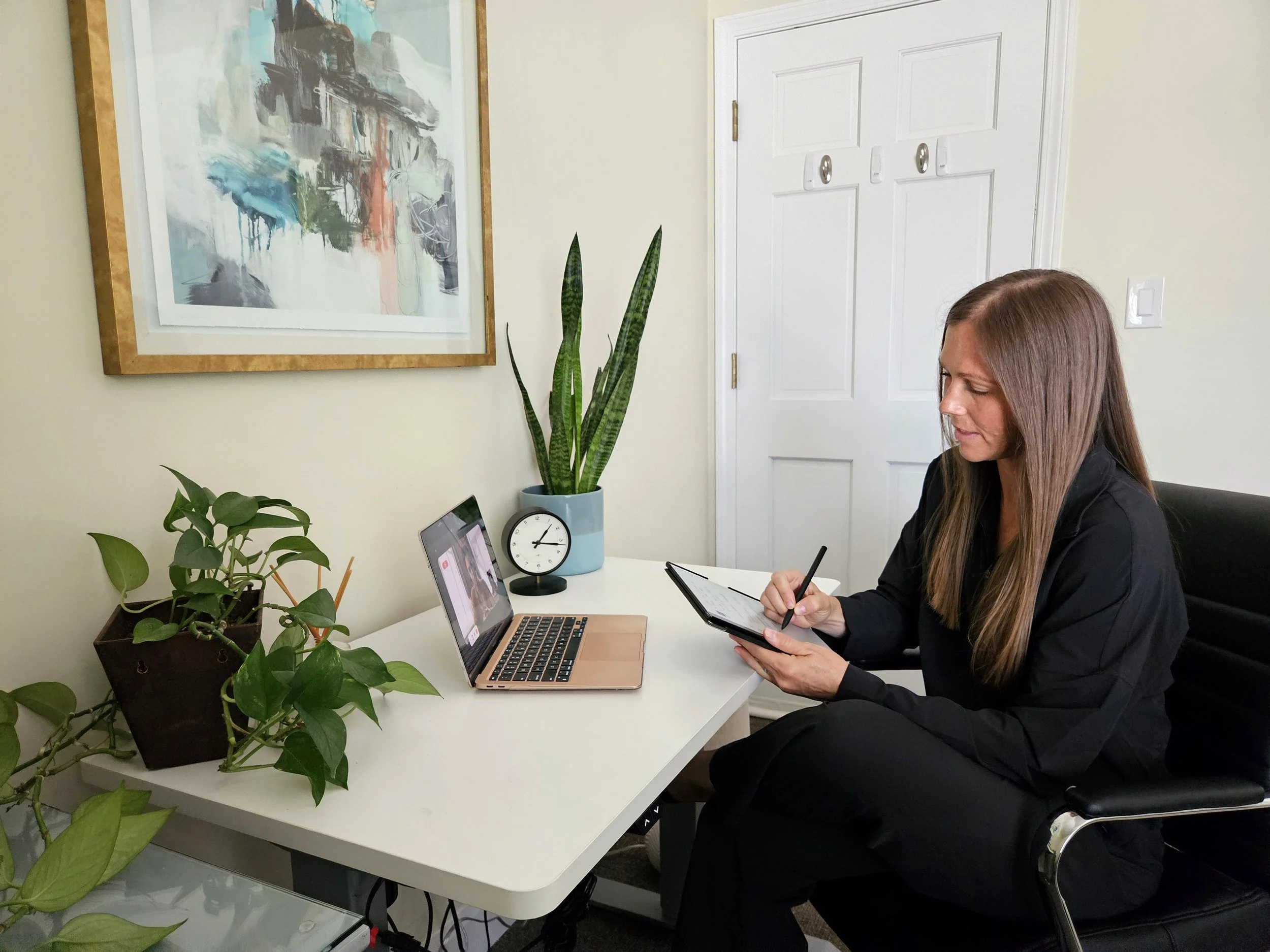 A woman sitting at a white desk in front of a laptop, writing in a notebook, with houseplants and a clock on the desk, an abstract painting on the wall, and a white door in the background.
