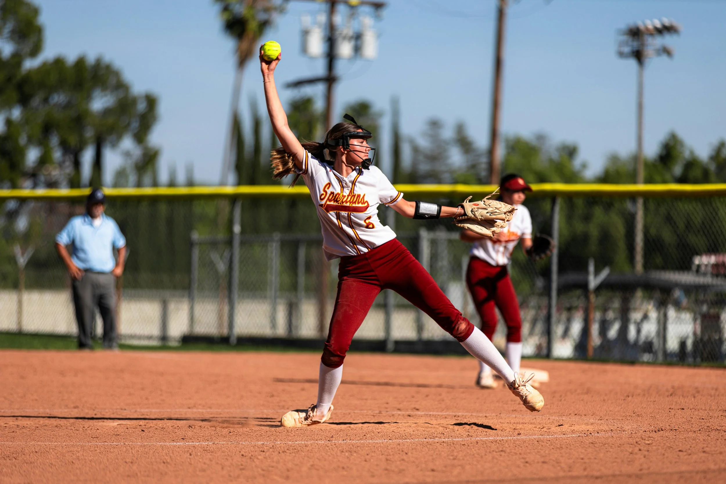 A female softball player in a white and red uniform with 'Spartans' written on it, is pitching a yellow softball on a baseball field. She is wearing glasses, a glove on her left hand, and a protective face mask. There are other team members and an umpire in the background.