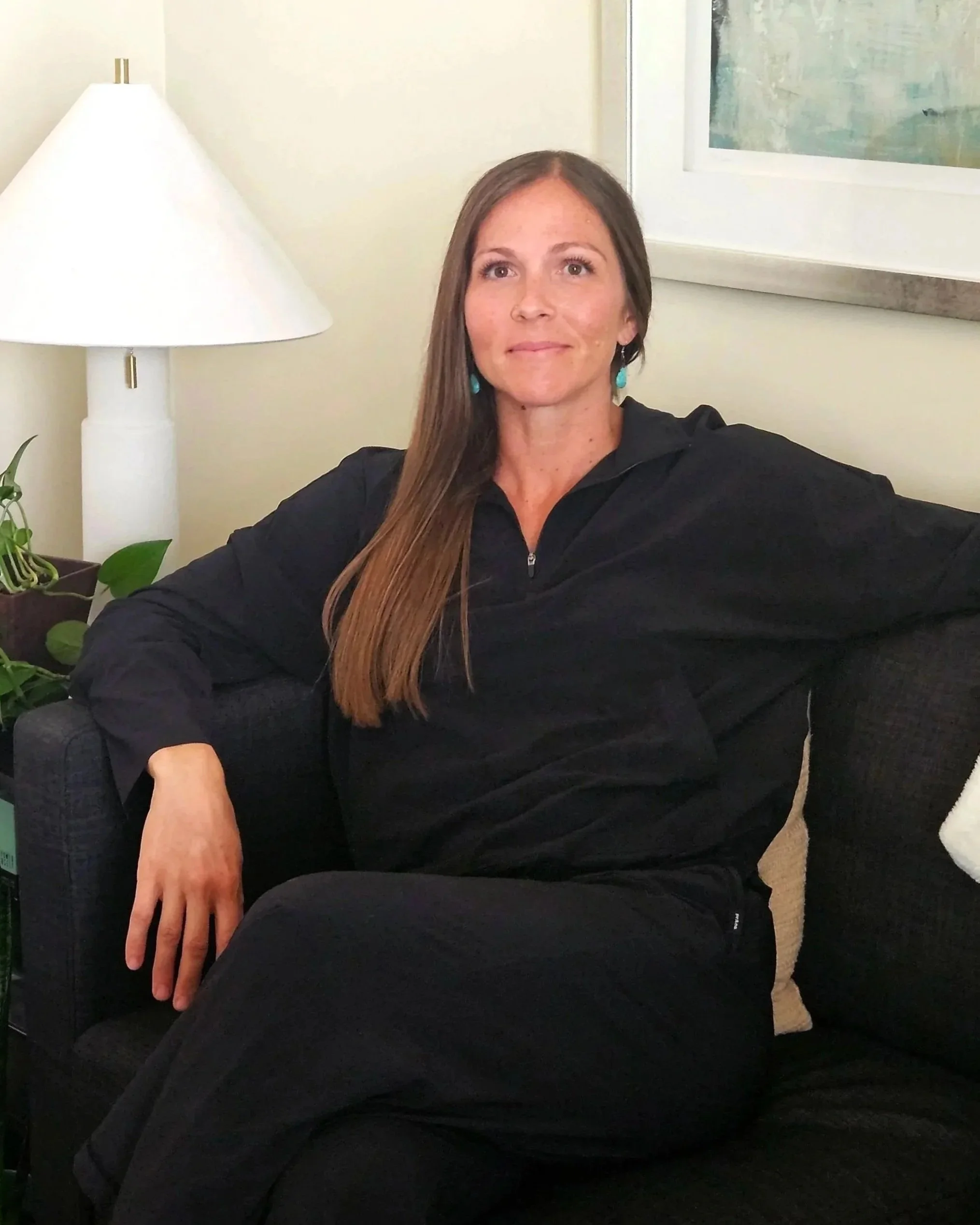 CEO and owner, Sonja, with long brown hair wearing a black top and turquoise earrings sitting on a dark sofa in a room with a white wall, a white lamp, and a plant.