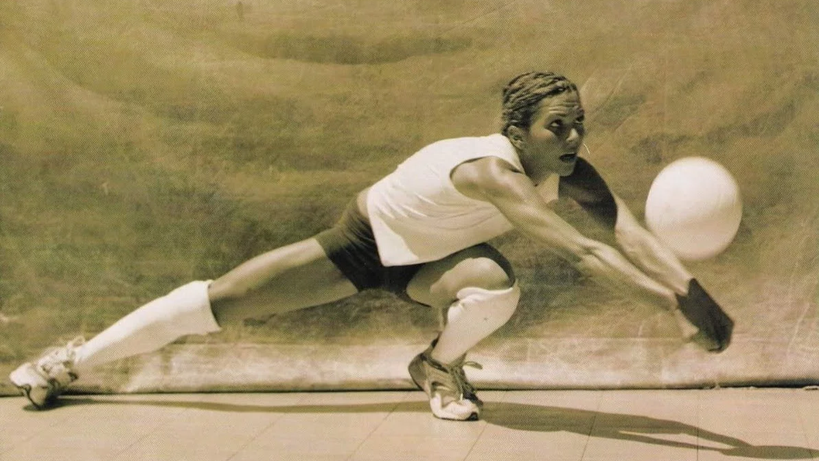 Female athlete in sports attire lunging to reach a volleyball on an indoor court.
