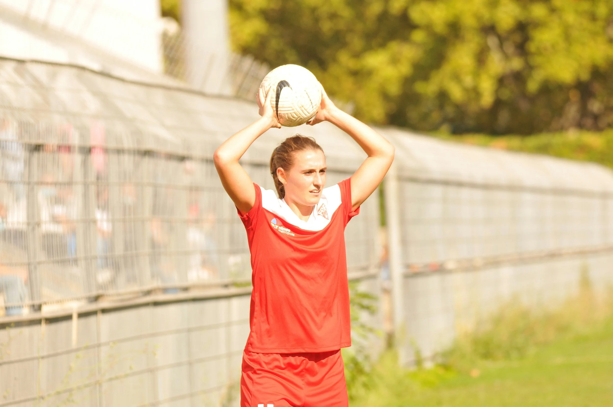 A woman in a red sports uniform holding a soccer ball above her head on a field near a fence and trees.
