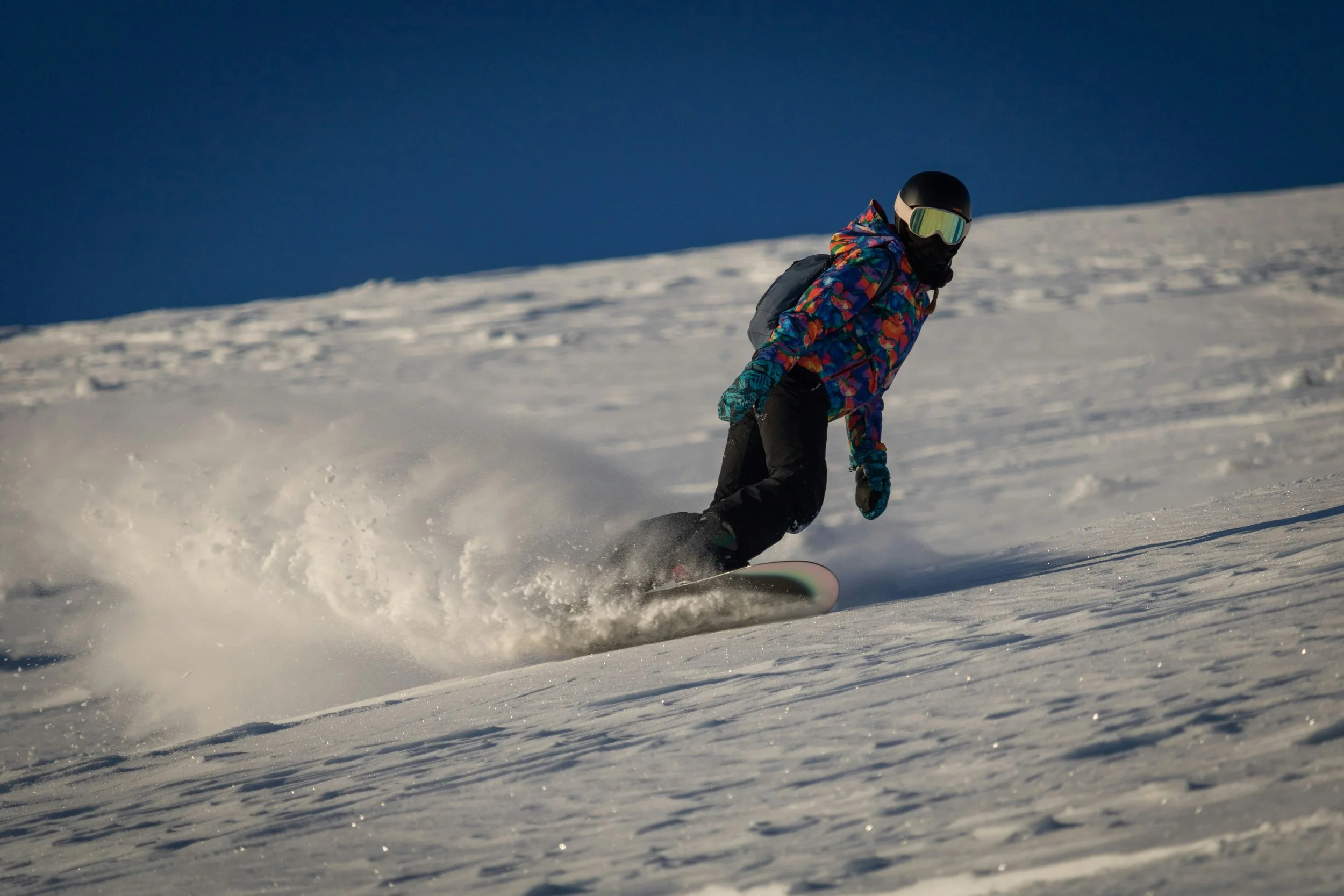A person snowboarding down a snowy slope during daytime, wearing a colorful jacket, black pants, a black helmet with goggles, and a backpack. The sky is clear and blue.