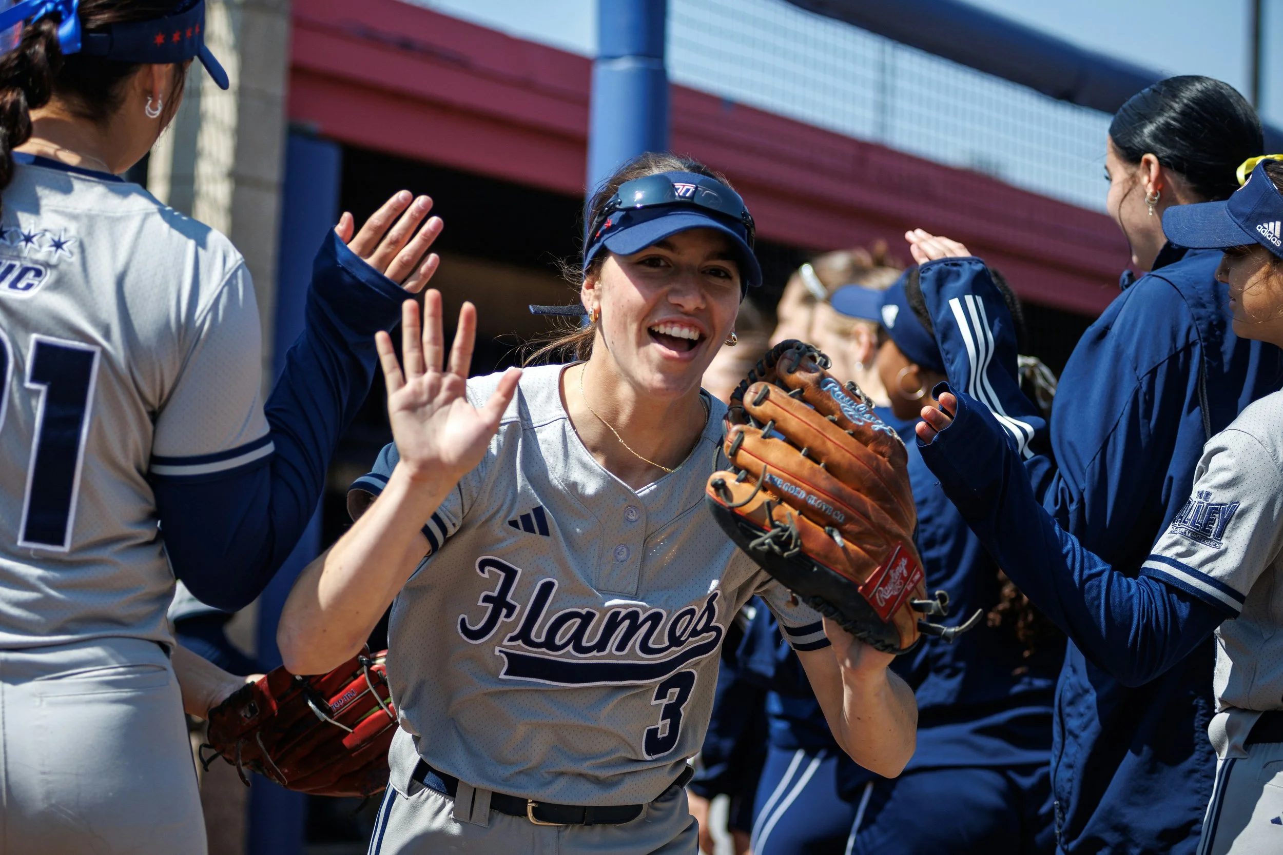 A group of female softball players celebrating on the field. One player is smiling at the camera, waving, and wearing a glove. The players are wearing uniforms with the team name and numbers visible, and some are wearing hats and visors. They appear to be in a good mood after a game.