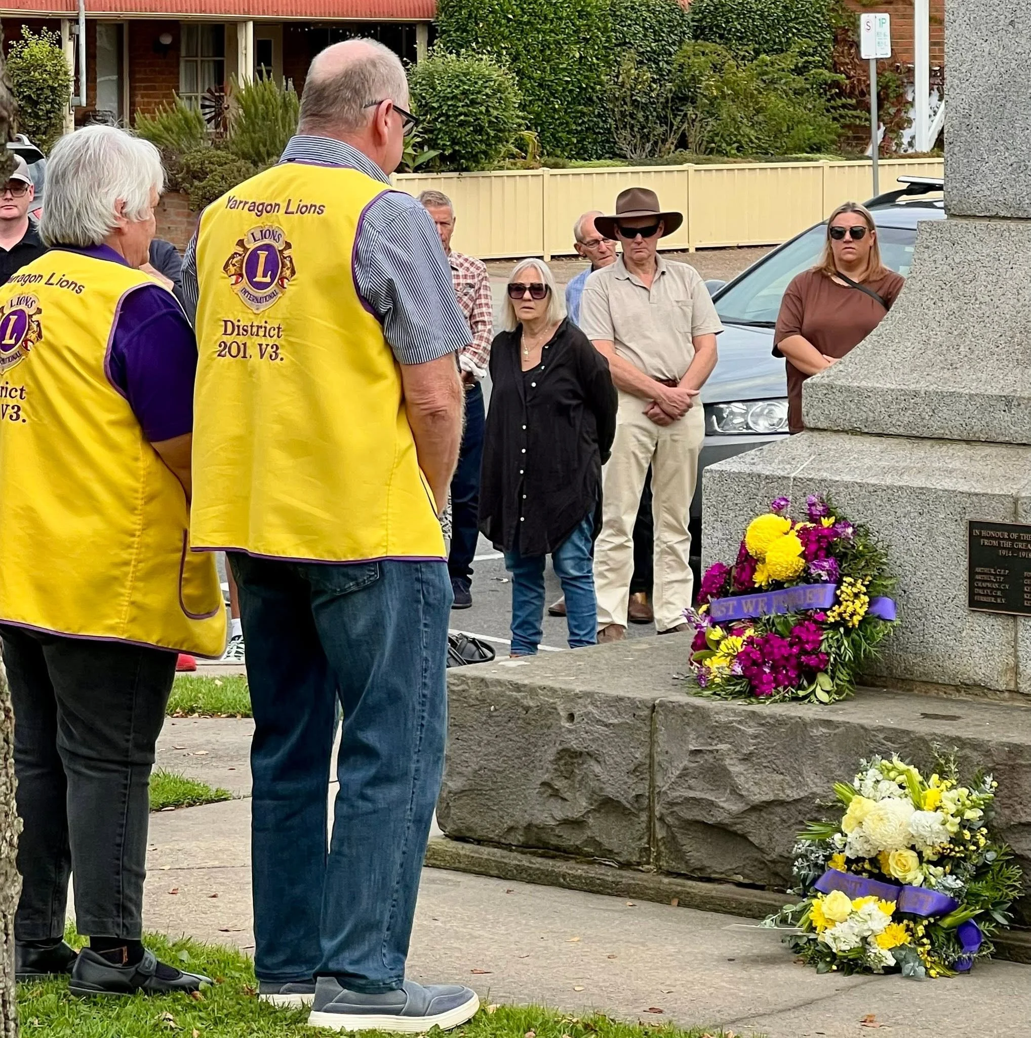 People gather around a memorial with flower wreaths at a remembrance event. Several attendees are wearing yellow vests with Lions Club insignia and some are wearing sunglasses or hats. The background features a brick house and a beige fence.