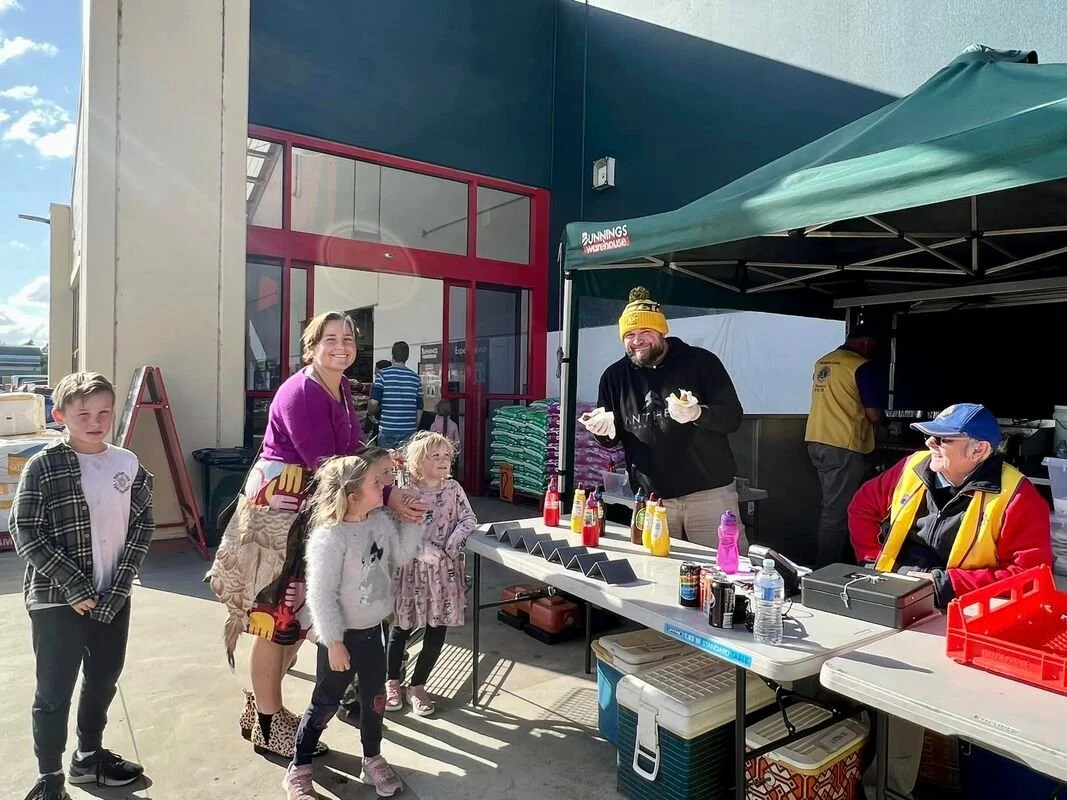 Children and adults standing in line at a food stall outside a building, with ketchup and mustard bottles on the table, and a man wearing a yellow beanie serving food.