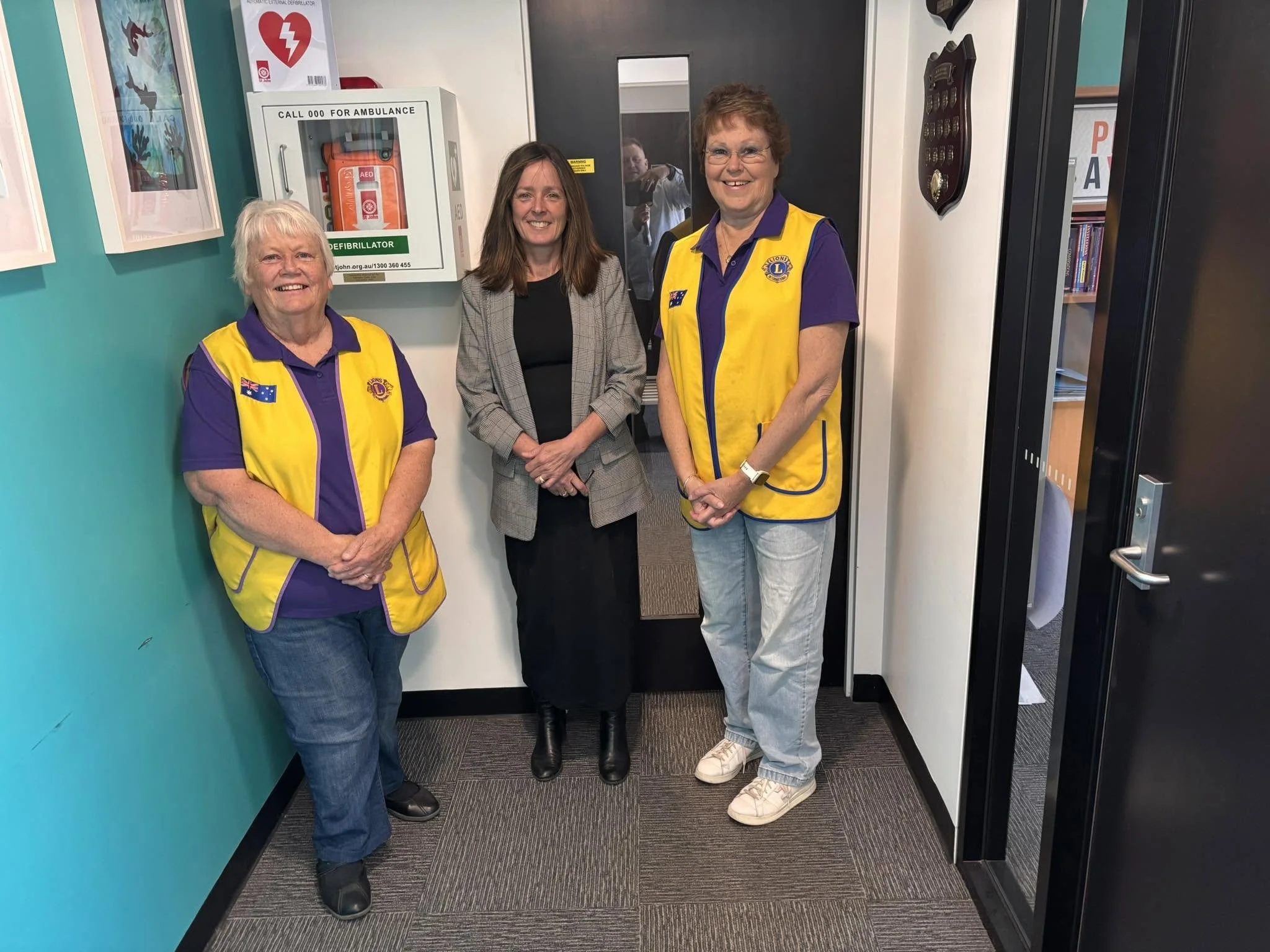 Three women standing indoors, smiling at the camera. Two women are wearing yellow and purple shirts with Lions Club logos, while the woman in the middle is dressed in business attire. Behind them is a gray door and some framed artwork on the wall.