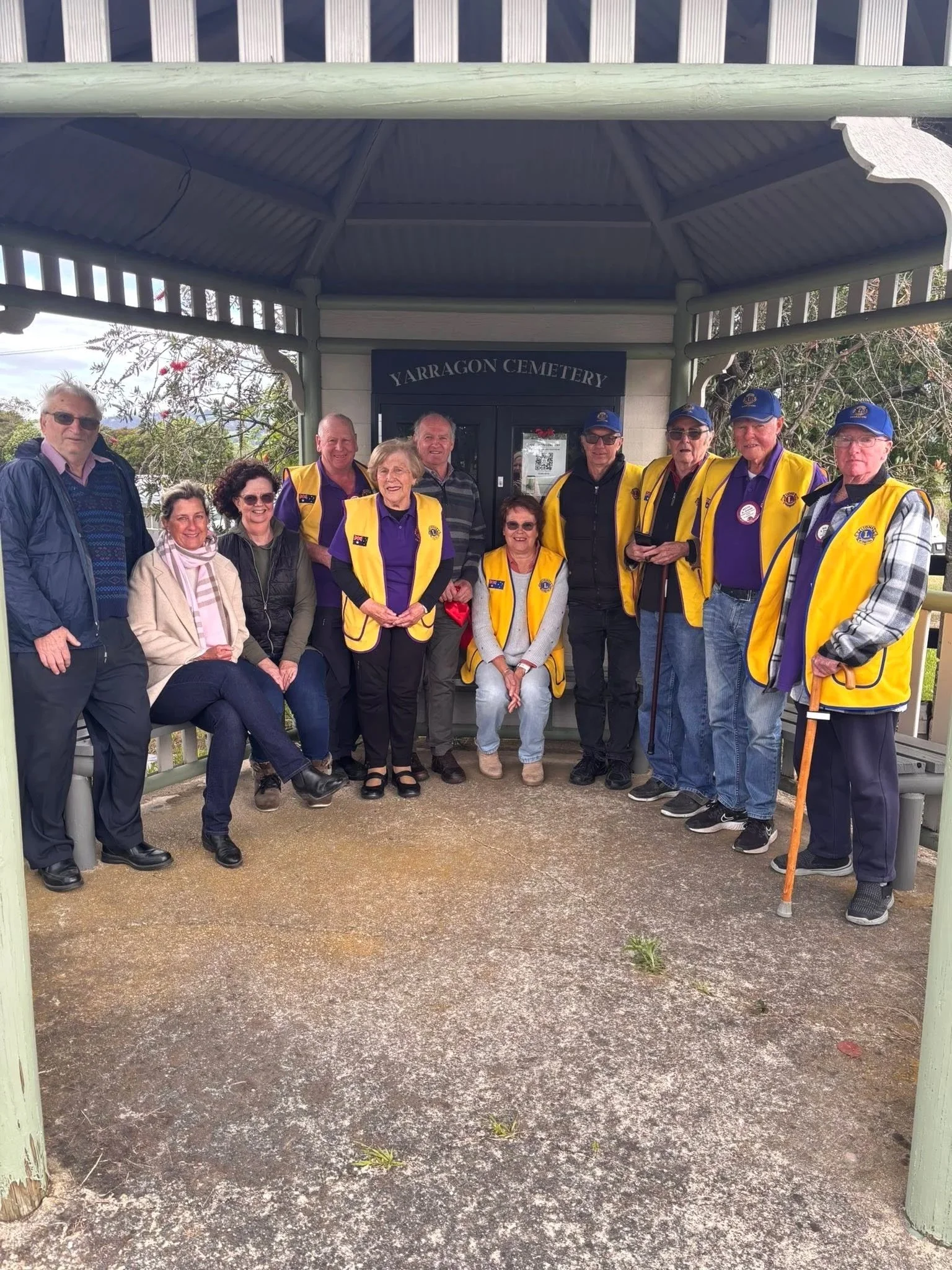 A group of people, mostly seniors, standing and sitting in front of a sign that says 'Yarragon Cemetery' under a gazebo. The group is wearing yellow vests and casual clothing, some with hats and sunglasses. One person has a cane.