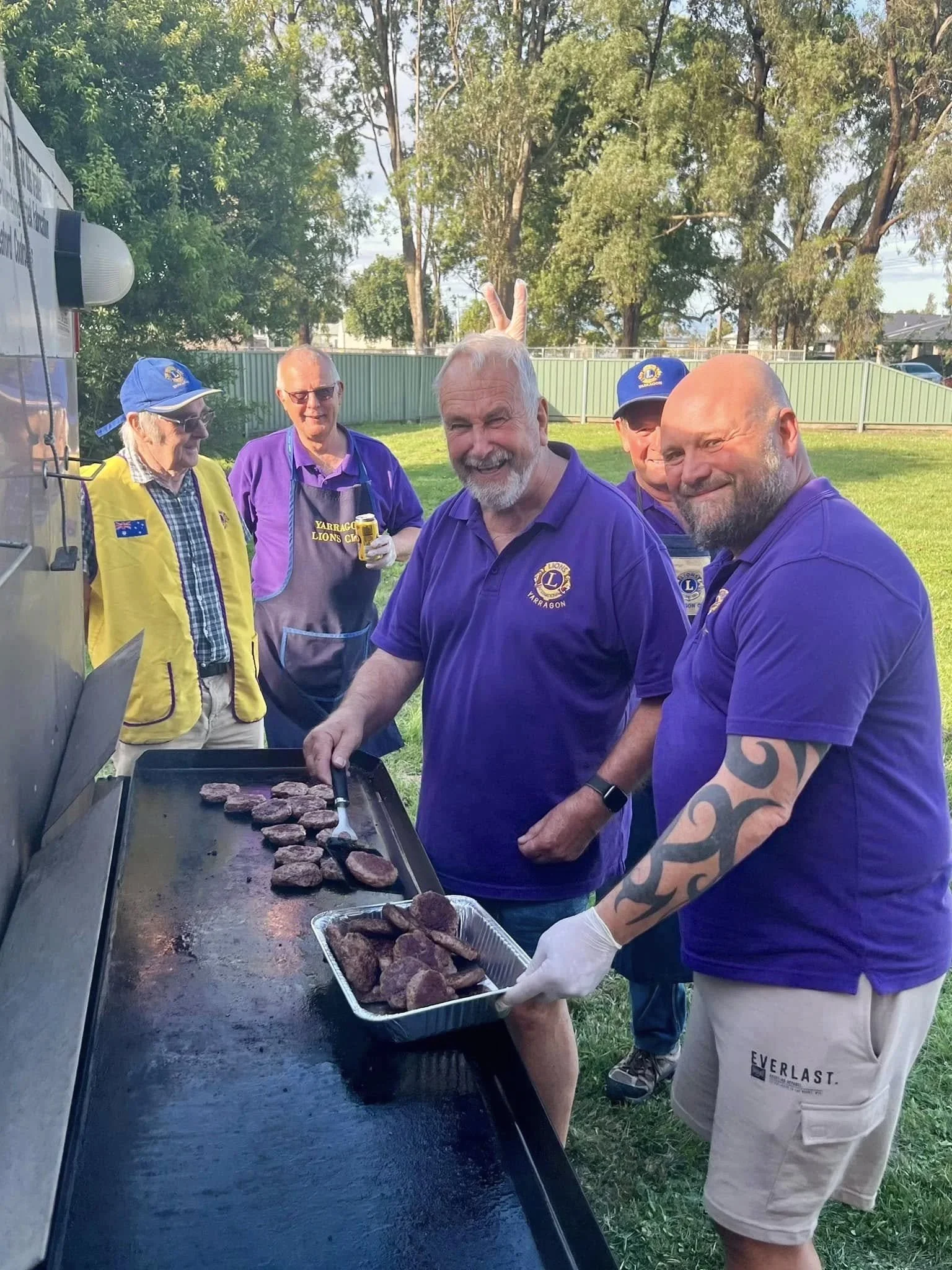 A group of five men at an outdoor barbecue, grilling and serving cooked meat, some smiling and one making a peace sign with his fingers.