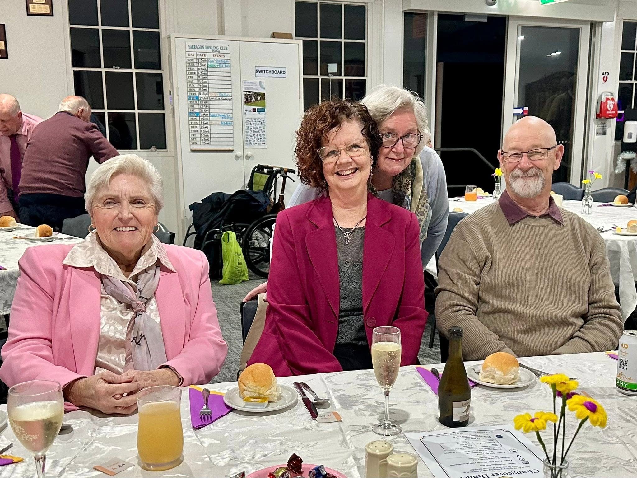 Four elderly adults, three women and one man, are gathered around a table at a social event, smiling at the camera. The table is set with plates, glasses, bread rolls, and small vases with yellow and purple flowers, and there are purple napkins and u