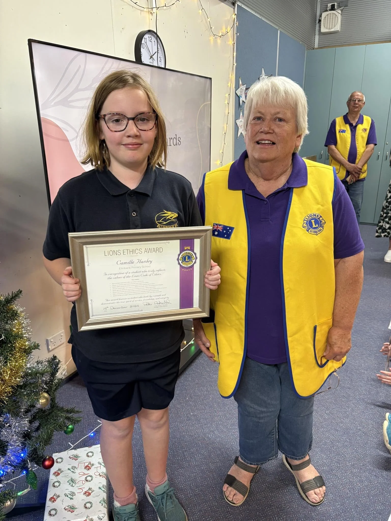 A girl holding an award certificate standing next to an older woman wearing a Lions Club vest, in a festive setting with Christmas decorations and a decorated tree.