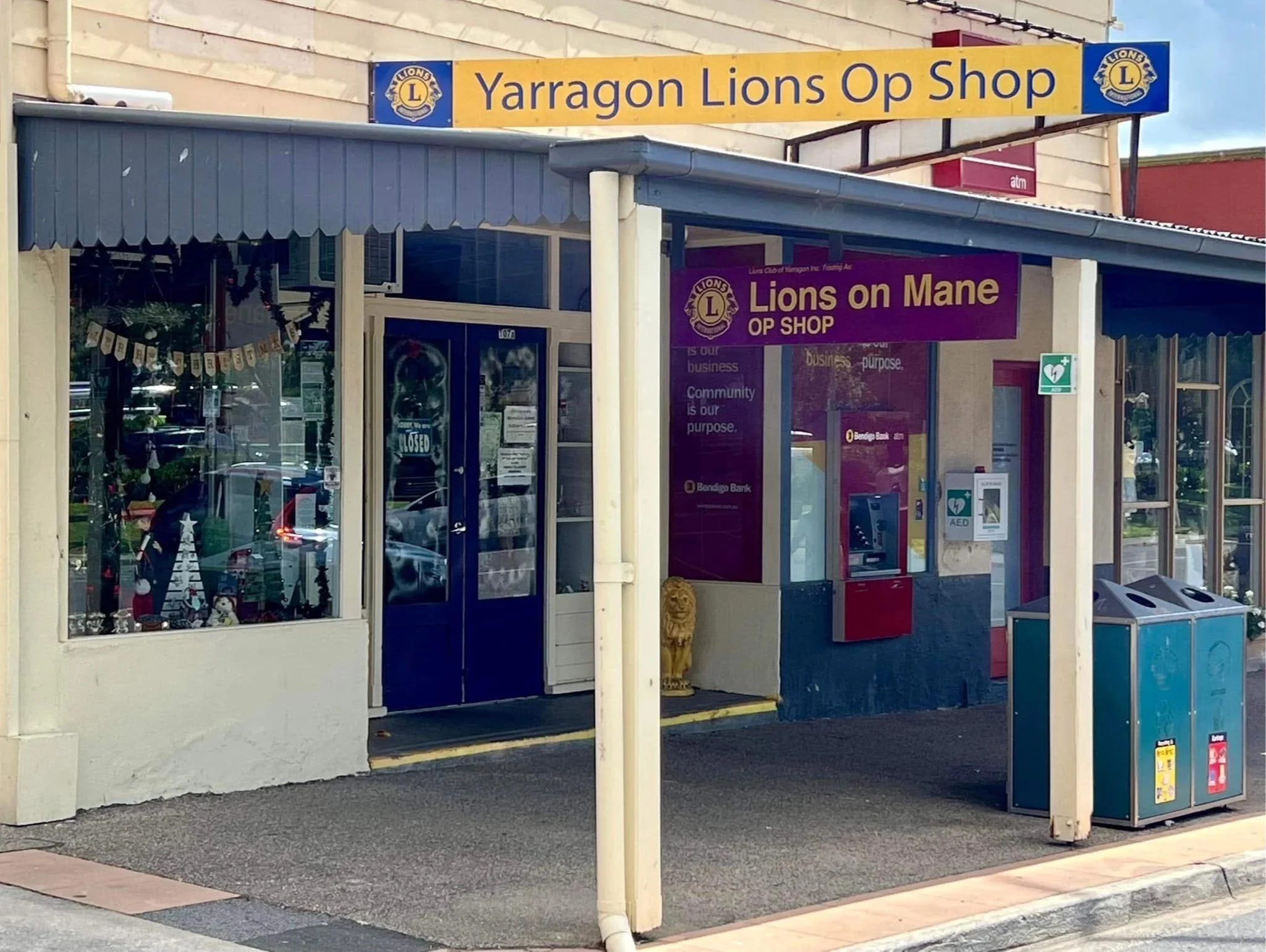 The image shows the front entrance of a small shop called 'Yarragon Lions Op Shop,' with a yellow sign and a blue Lions Club logo. There are large windows displaying various items inside, and a purple sign to the right that reads 'Lions on Mane OP SHOP.' Outside, there's a red donation bin and some signs at sidewalk level. The building has a white exterior and a blue awning above the entrance.