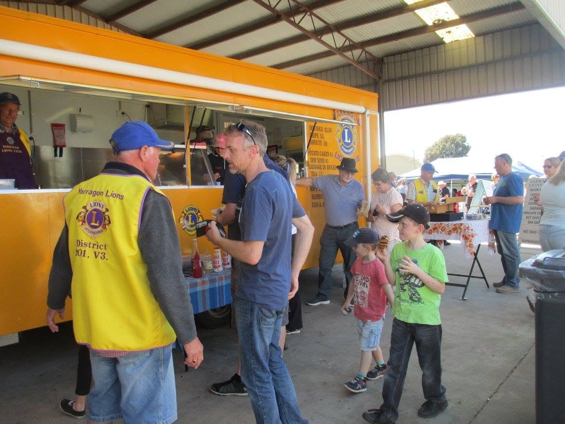 People standing in line at a food truck with a yellow exterior, serving food at an outdoor event under a large metal pavilion. Some individuals wear bright yellow vests with the Lions Club logo.