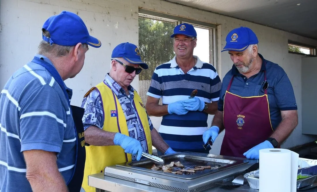 Four men wearing blue hats and gloves are gathered around a grill cooking meat, outdoors.