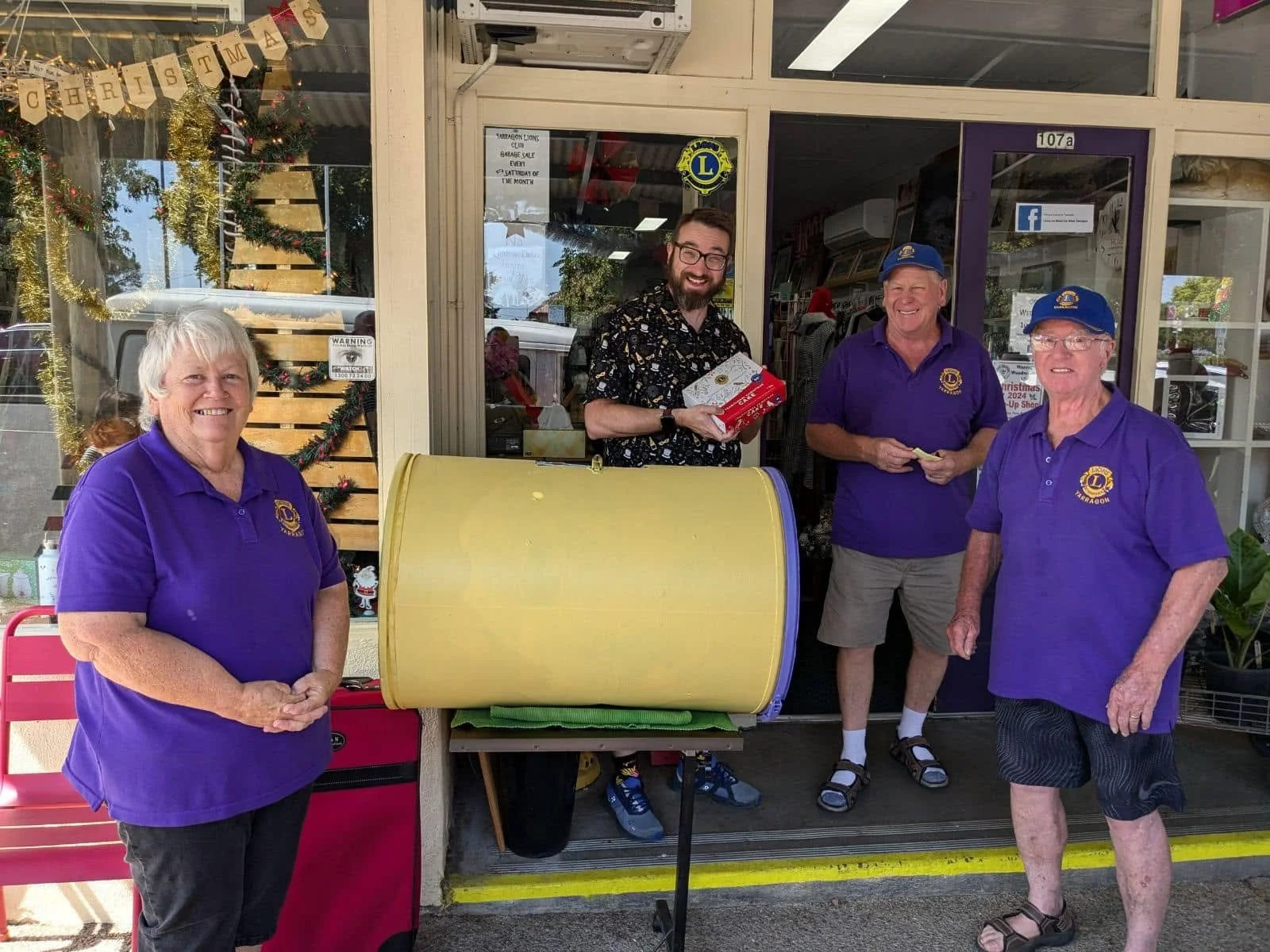 Four elderly people and a younger man standing in front of a store, with a large yellow donation barrel, Christmas decorations, and a red chair visible.