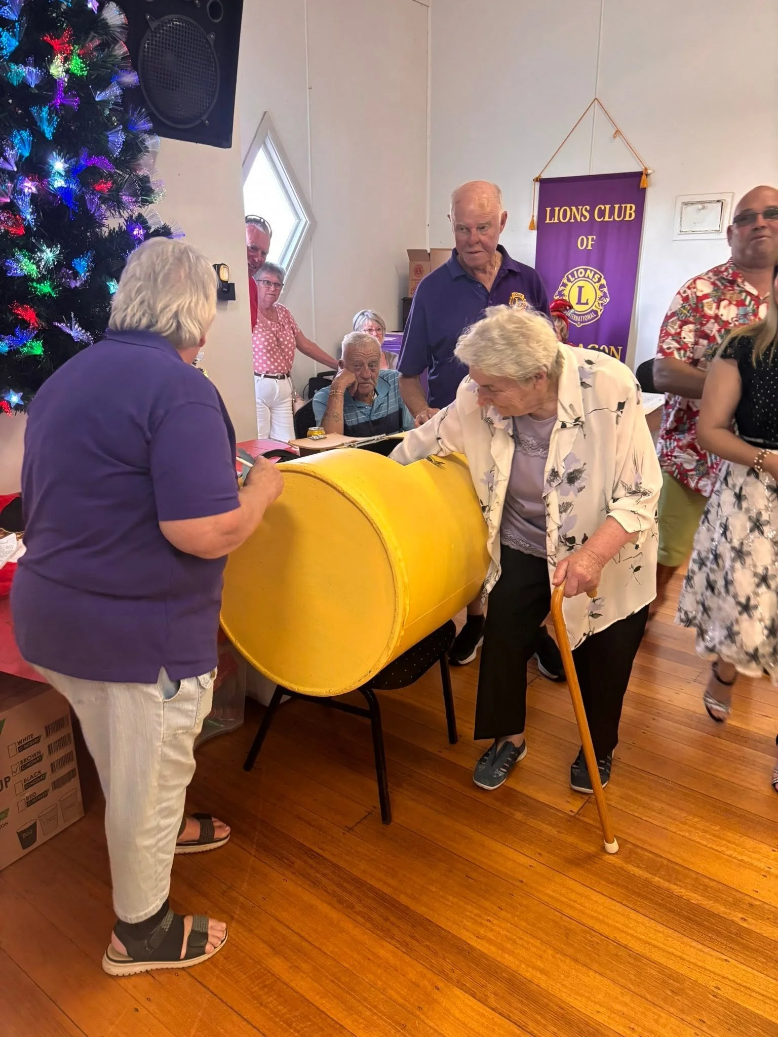 Group of elderly people participating in a raffle at a Lions Club event, with a Christmas tree and colorful decorations in the background.