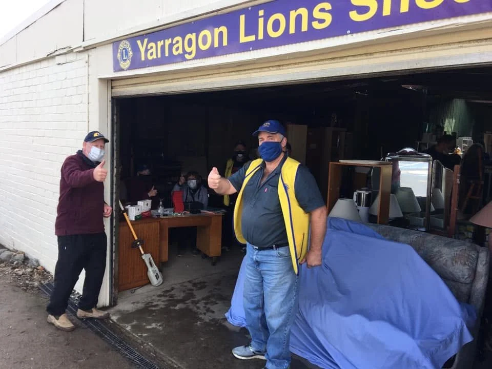 People standing outside a store called Yarragon Lions Shop, wearing masks and giving thumbs-up gestures, with furniture inside visible through the open garage door.