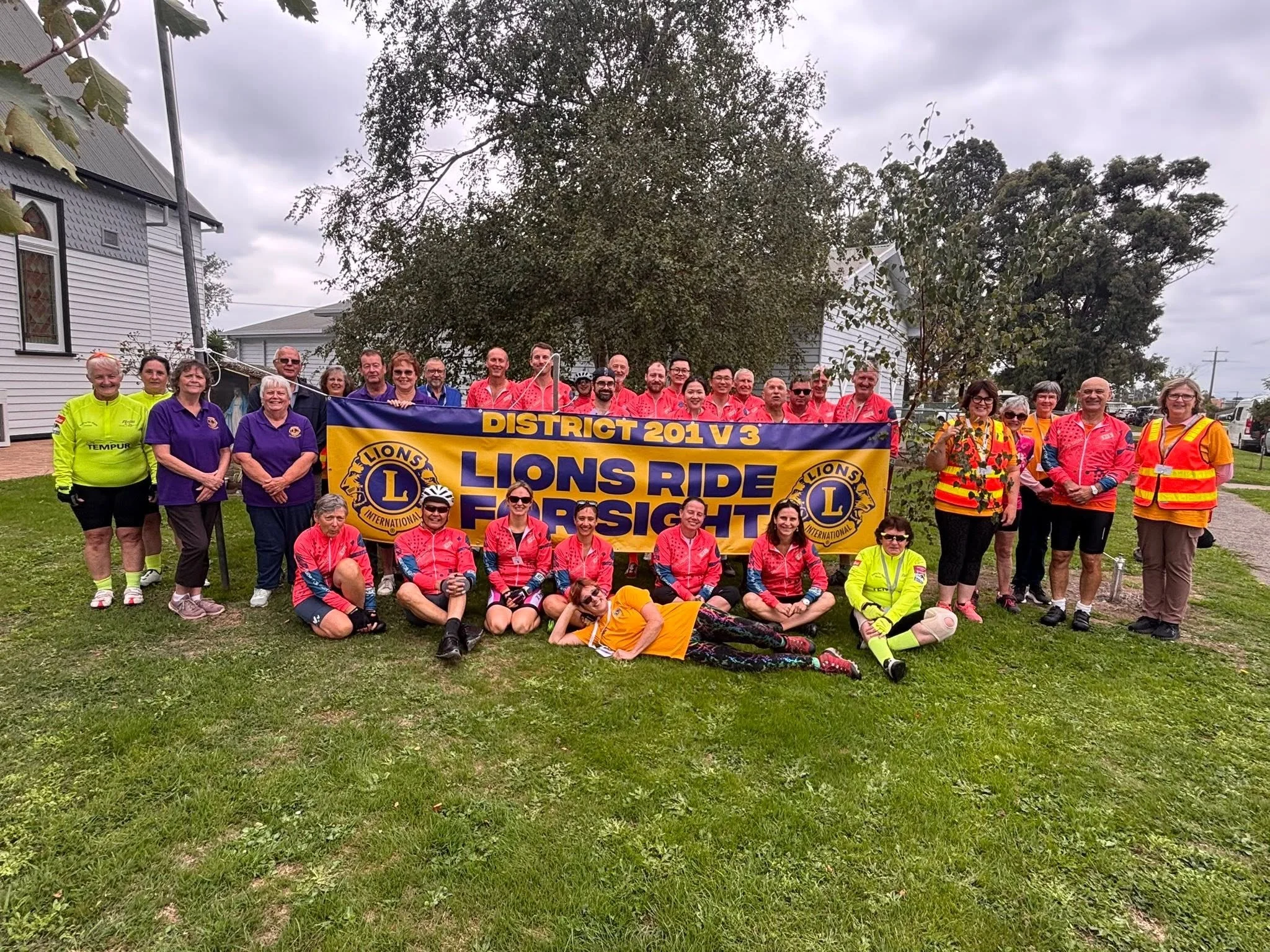 A group of people, mostly wearing red shirts, holding a blue and yellow banner that says "Lions Ride for Sight" with the Lions Club logo, standing outdoors on grass with trees and houses in the background under an overcast sky.
