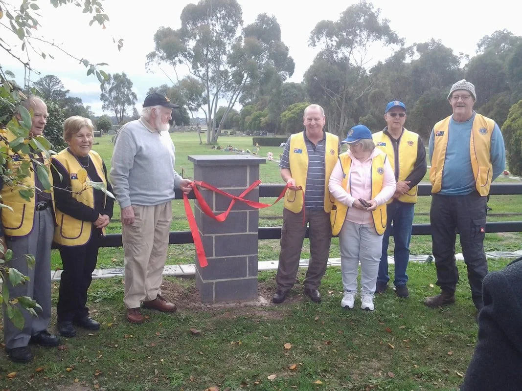 Group of people, some wearing yellow vests, gathered for a memorial or unveiling ceremony outdoors in a park, with a small stone marker and red ribbon.