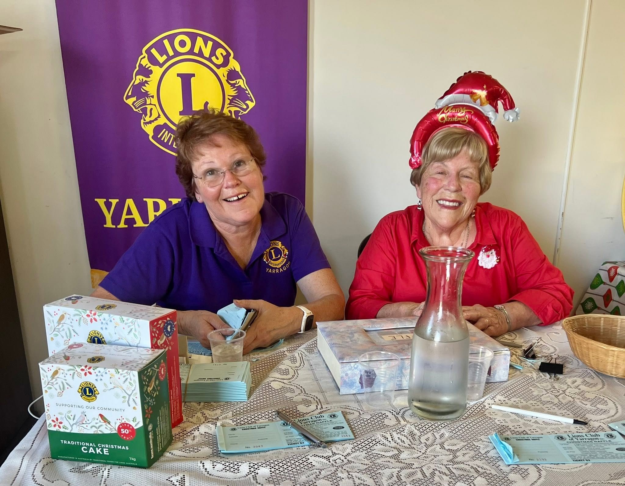 Two women are sitting at a table during an event, smiling at the camera. One woman is wearing a purple shirt with a Lions Club logo, and the other is in a red shirt with a Christmas themed balloon headpiece. The table has various items, including pamphlets, a clear pitcher of water, papers, and a decorated box. A purple Lions Club banner hangs in the background.