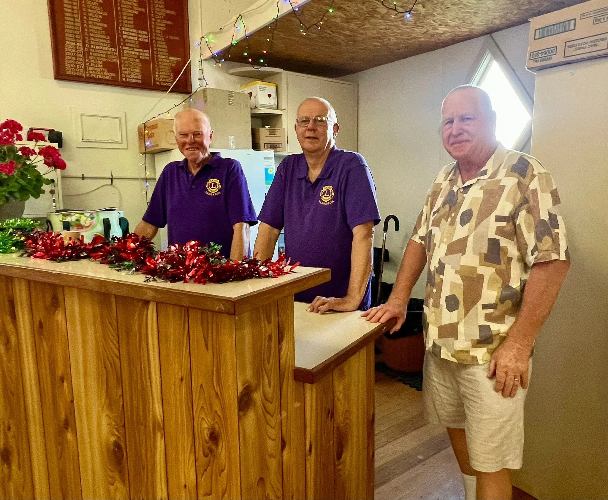 Three elderly men standing behind a wooden counter decorated with red tinsel and greenery, in a room with Christmas lights, a yellow flowered plant, and various boxes and cabinets visible in the background.