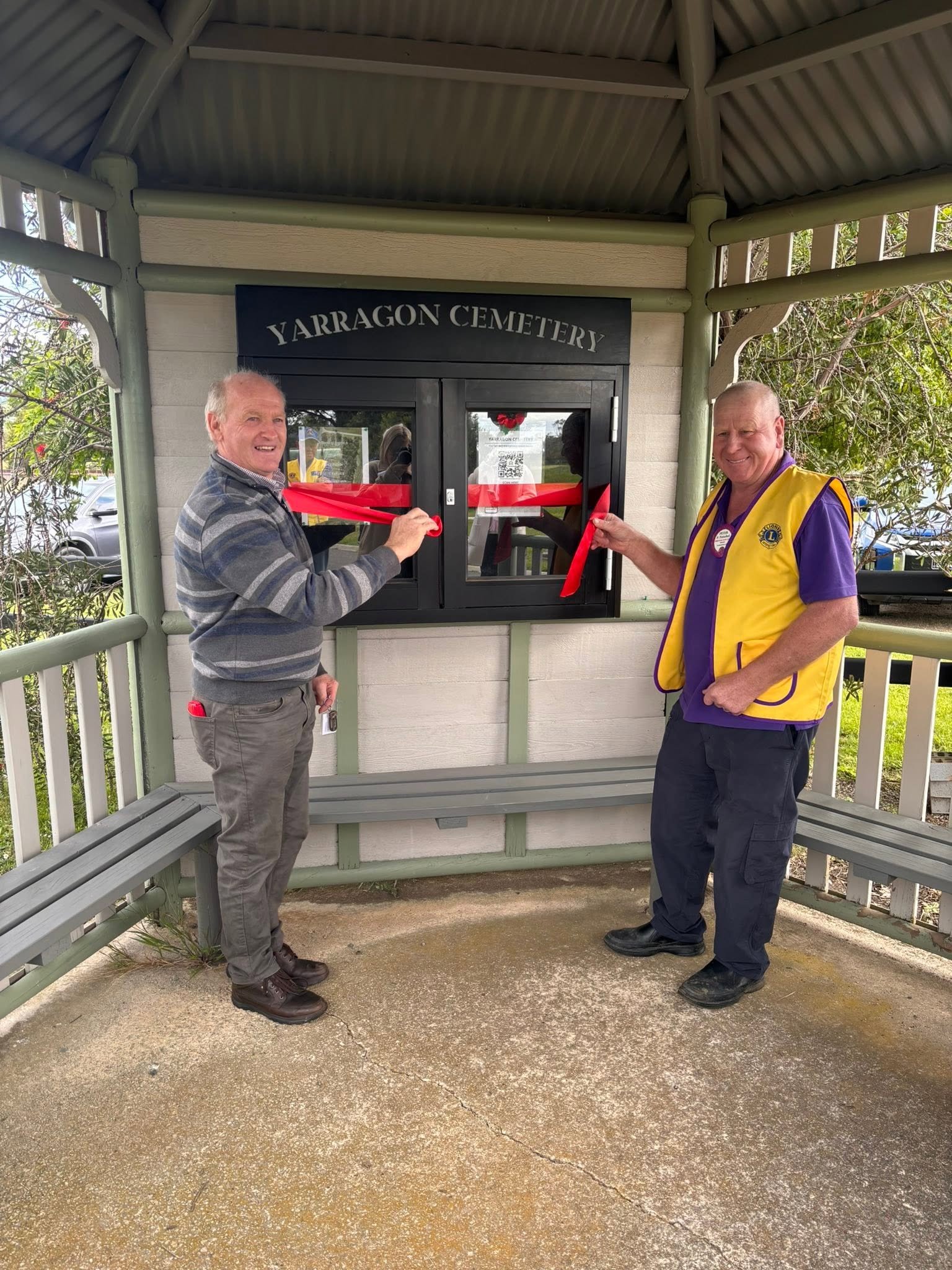 Two men stand under a gazebo, cutting a red ribbon in front of a sign that reads 'Yarragon Cemetery,' celebrating its opening or dedication.