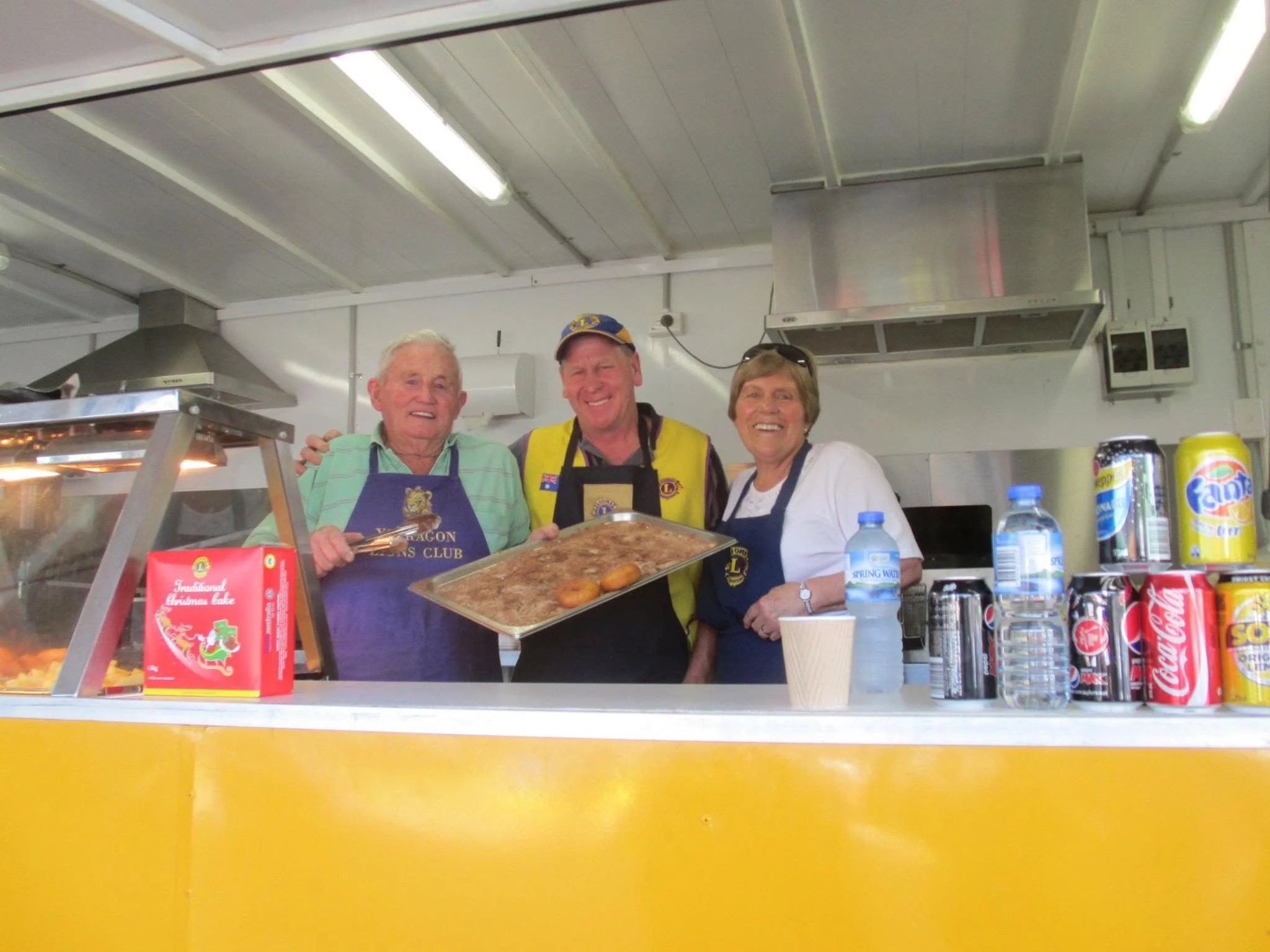 Three seniors smiling in a kitchen, holding food trays, with drinks and snacks on the counter in front of them.