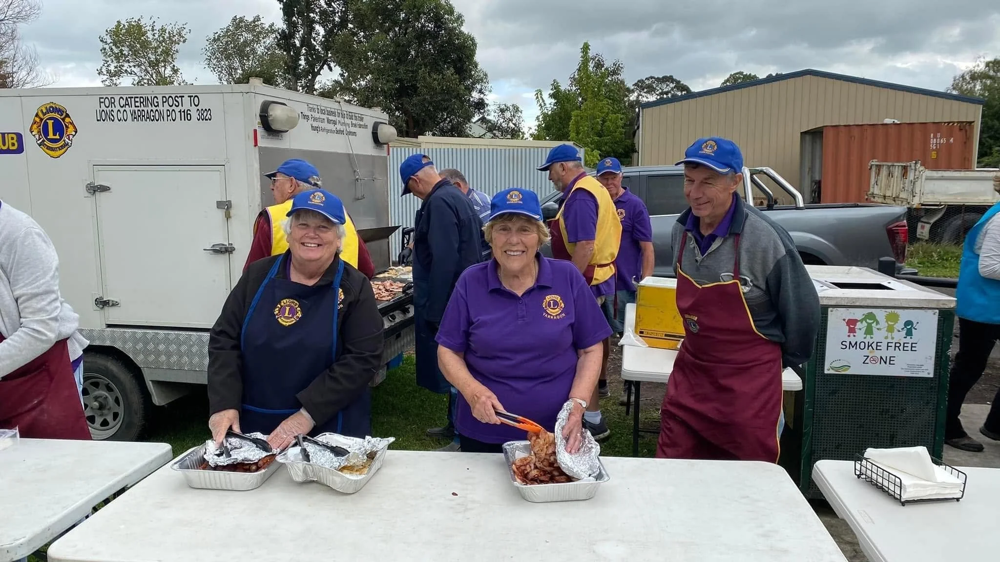A group of people, mostly seniors, are gathered outdoors at a barbecue event. They are wearing matching blue hats and some are wearing aprons with the Lions Club logo. There are tables with food, and a catering truck in the background. Several individuals are smiling and preparing or serving food, with a cloudy sky overhead.
