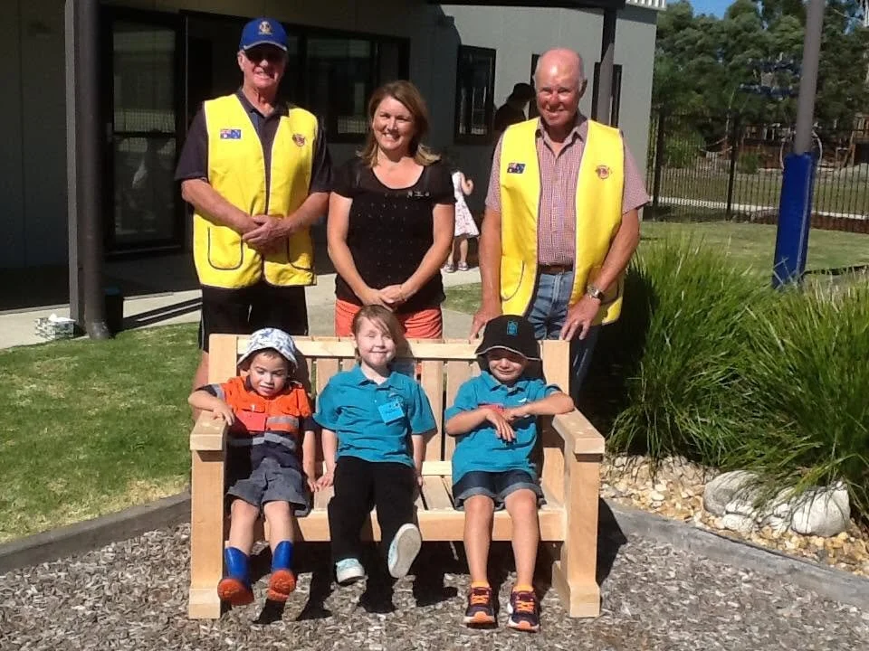 Group of six people outdoors, three adults standing behind three children sitting on a wooden bench. The adults are wearing yellow safety vests, and the children are in casual clothes, with one girl in an orange and gray uniform and the boys in blue 