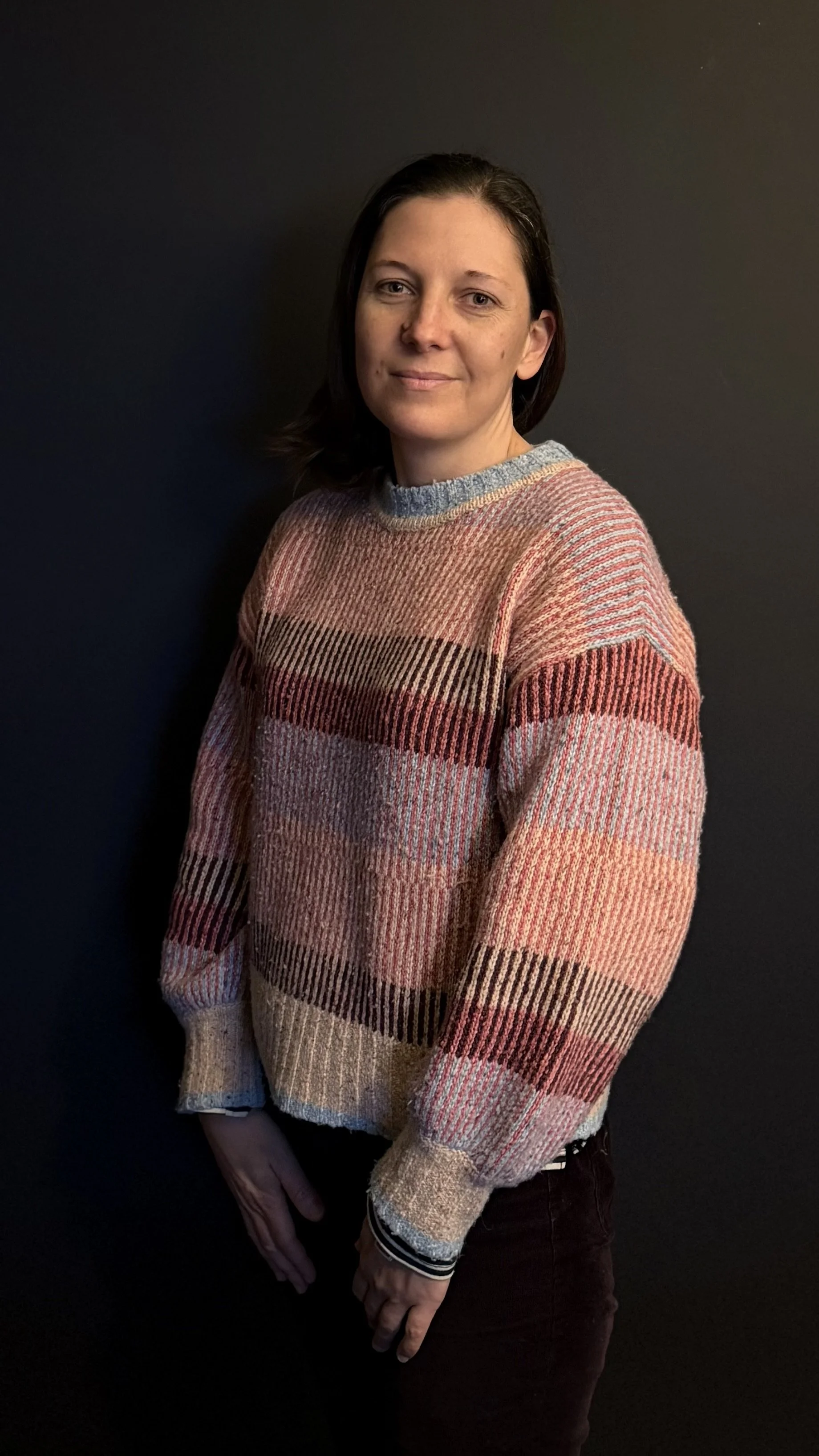 A woman with medium-length dark hair wearing a plaid sweater in shades of pink, beige, and brown, standing against a dark background.