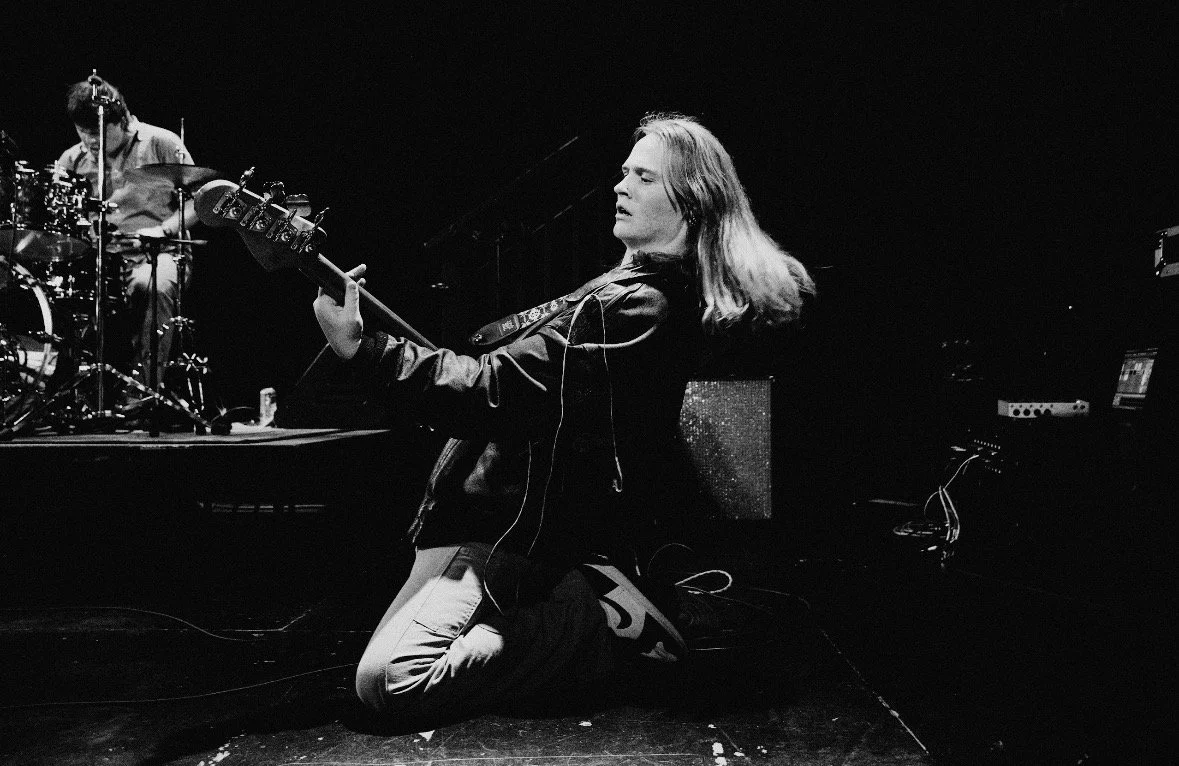 A male musician with long hair kneeling on stage, playing an electric bass guitar, with a drummer in the background.