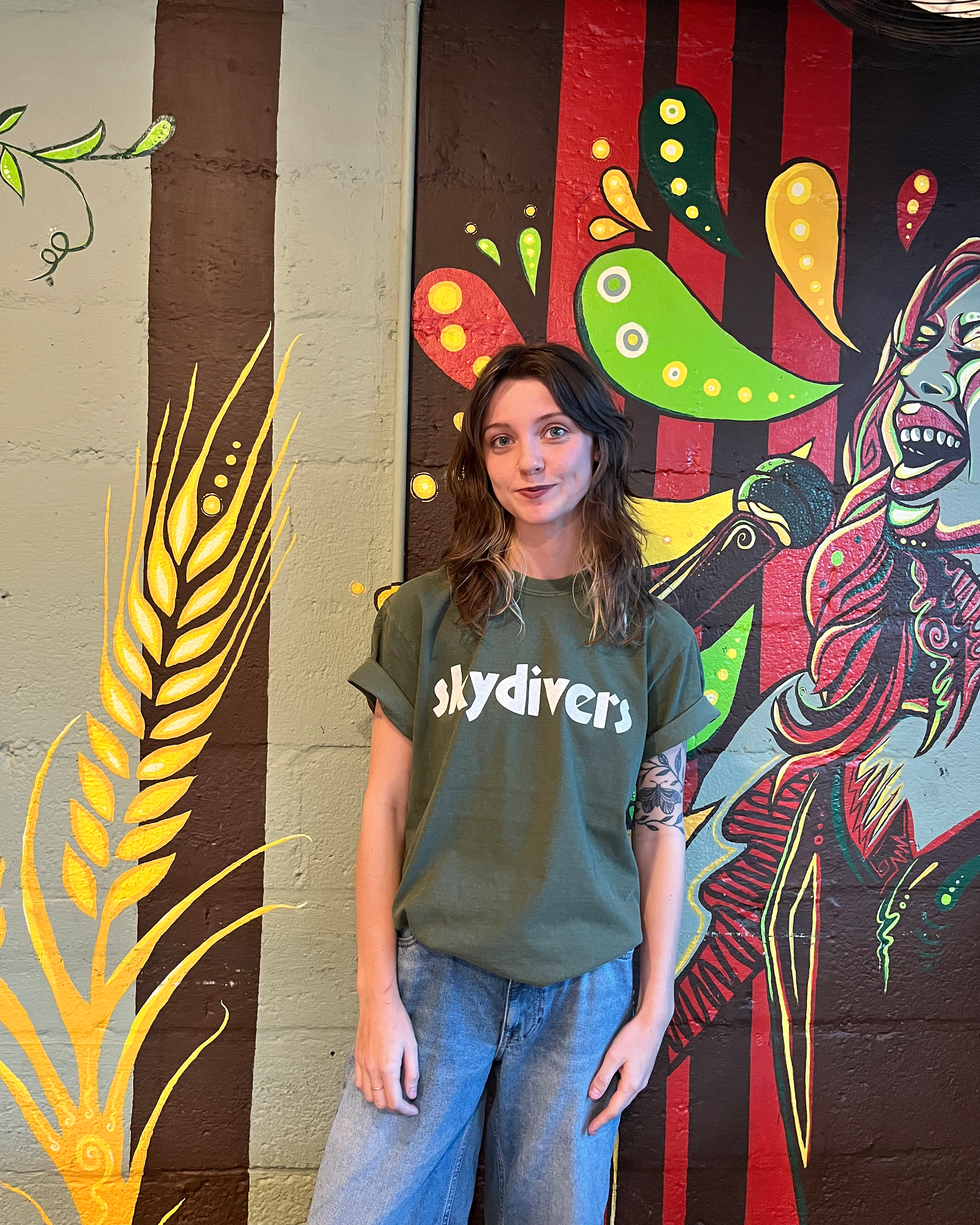 A young woman with shoulder-length brown hair and a nose piercing standing in front of a colorful mural painted on a wall. The mural features a green, yellow, and red abstract design with a face on the right side.