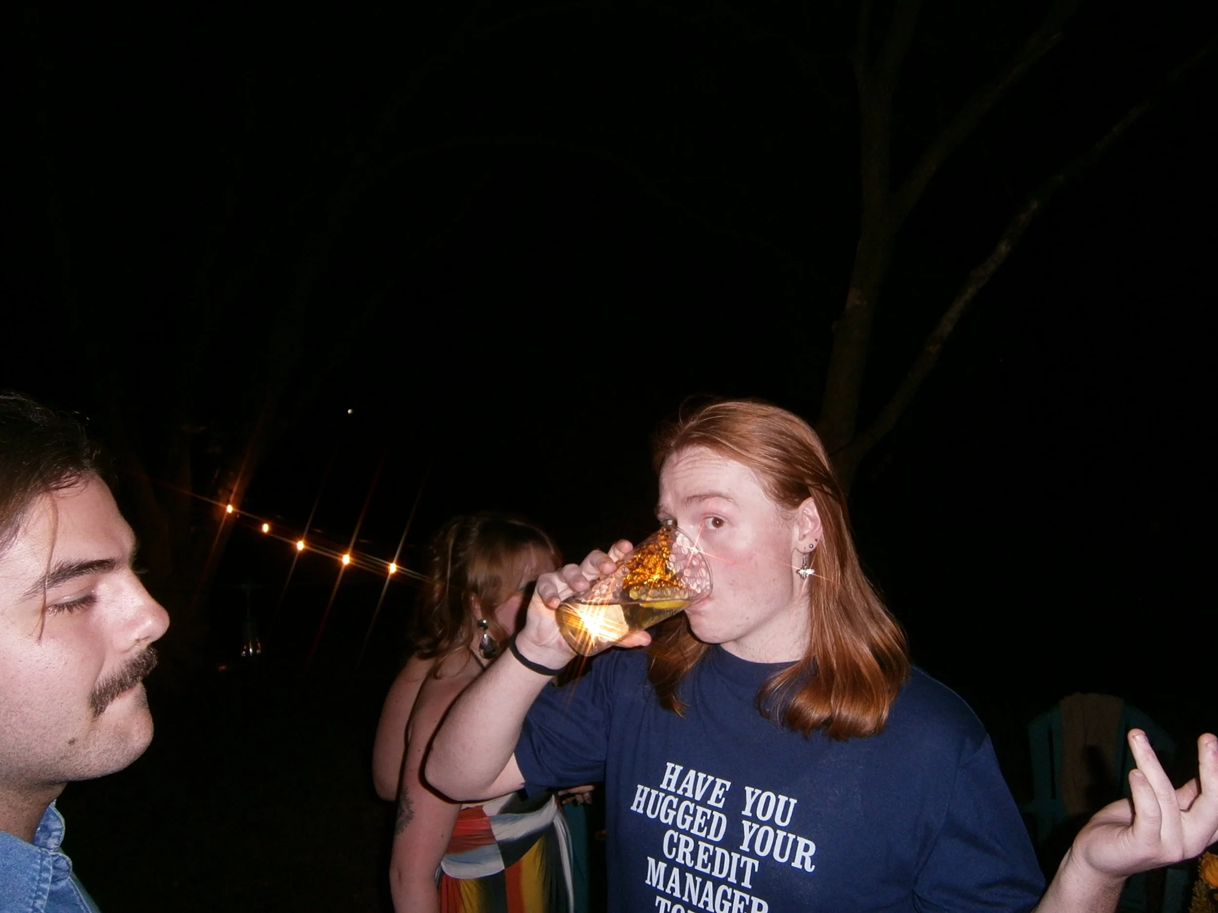 A group of people at night, with string lights in the background. A woman with red hair is drinking from a glass, and a man is on the left side of the image.
