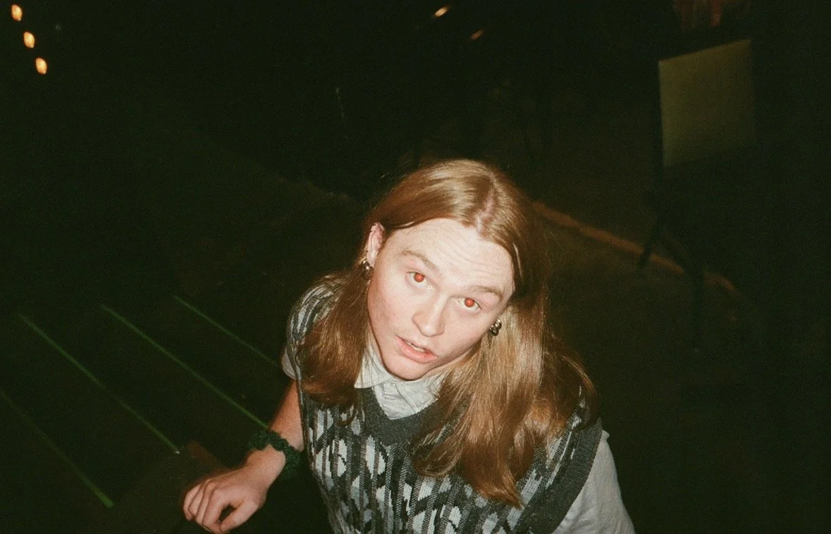 A young woman with long red hair looking up at the camera, wearing a patterned black and gray sweater over a collared shirt, in a dimly lit setting.