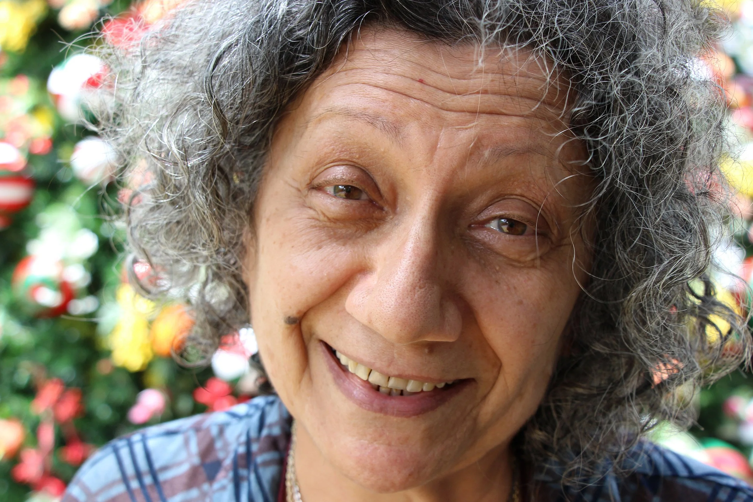 Close-up of a smiling elderly woman with curly gray hair, outdoors in front of a Christmas tree with colorful ornaments.