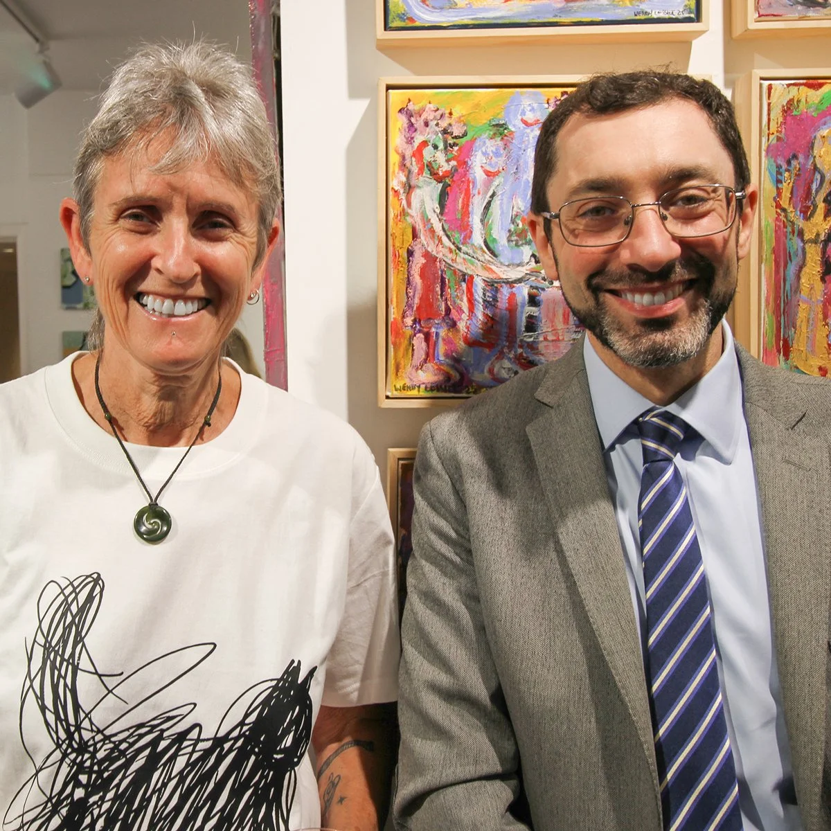 A smiling woman with short gray hair wearing a white t-shirt with black abstract art and a necklace with a green spiral pendant, standing next to a smiling man with glasses, dark hair, and a beard wearing a gray suit and striped tie. They are in an art gallery with colorful abstract paintings on the wall behind them.