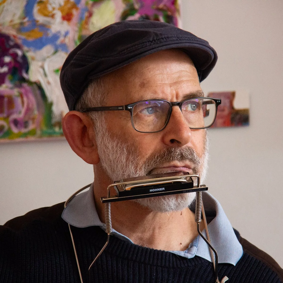 A man with gray hair, a beard, and glasses, playing a harmonica, wearing a cap and a sweater, with colorful abstract artwork in the background.