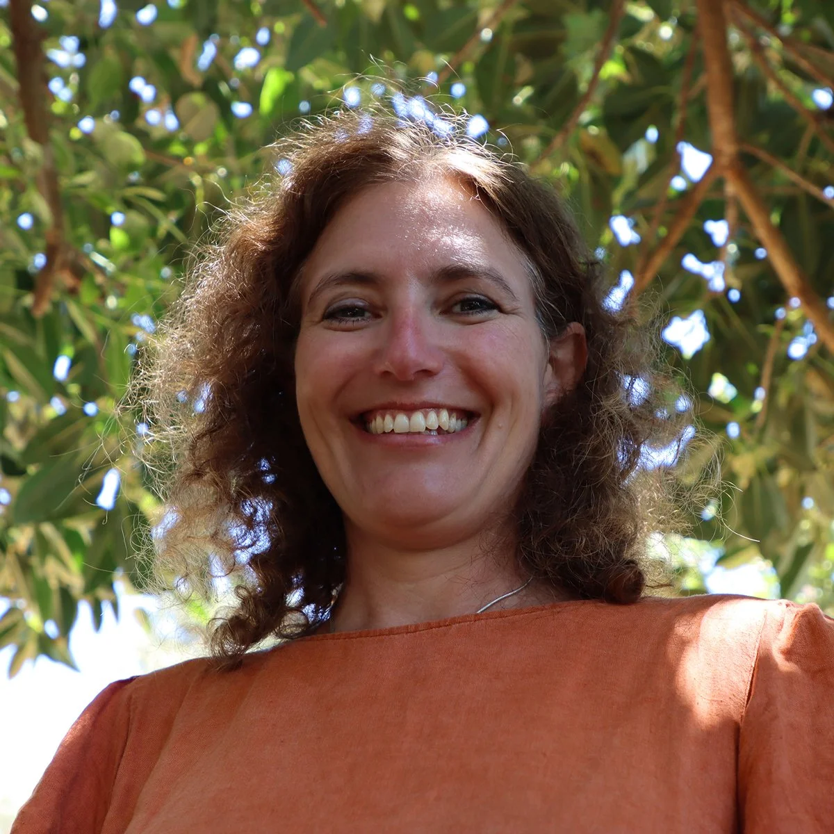 Smiling woman with curly brown hair standing outdoors in front of green leafy tree, wearing an orange top.
